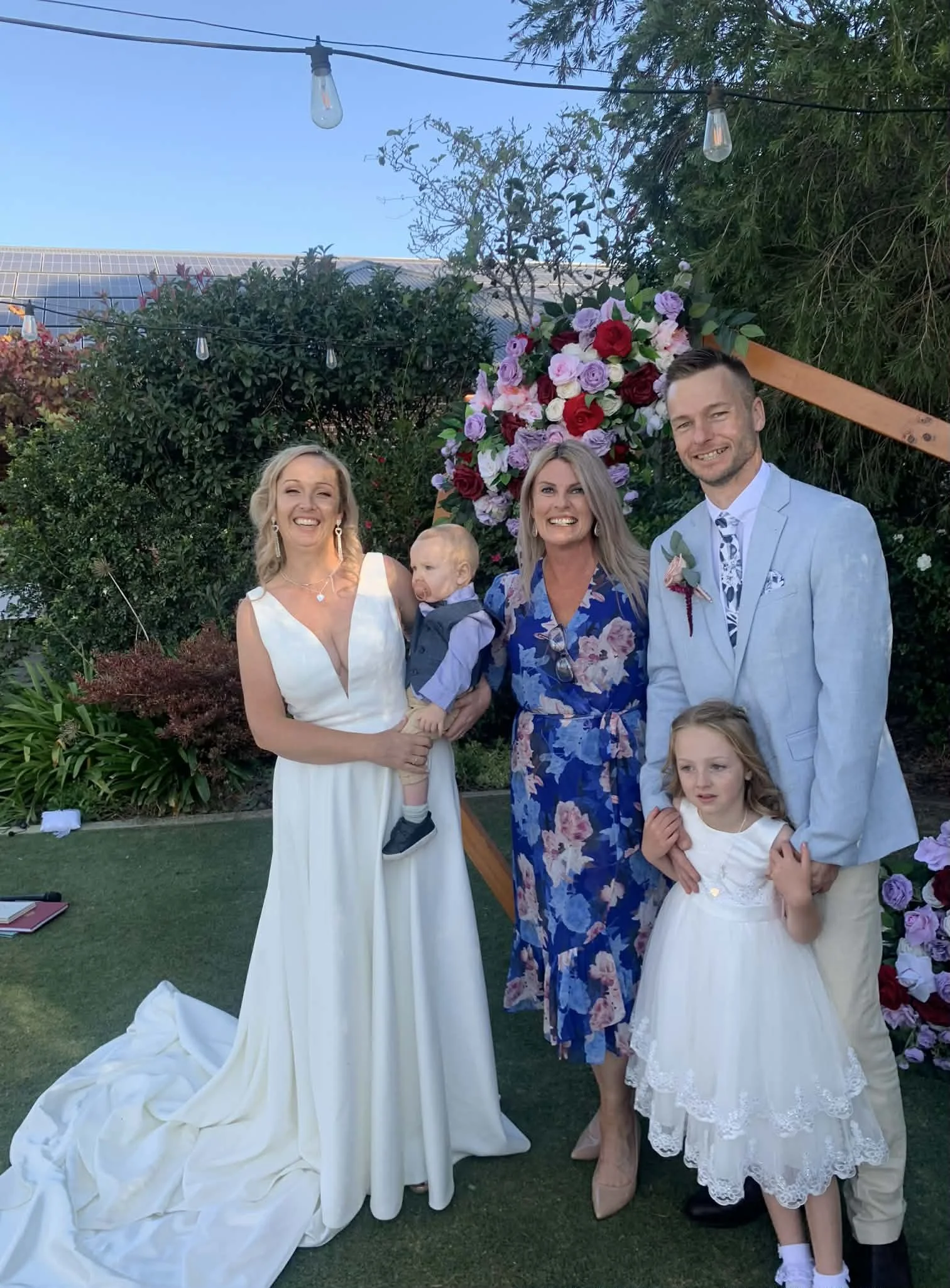 A group of five people and a child posing at a wedding ceremony outdoors, with a floral backdrop behind them. The woman on the left is wearing a white gown, and a man on the right is in a light suit. The woman in the middle is wearing a blue floral dress, and the girl in the dress is standing in front of the man. The boy is being held by the woman in the white gown.
