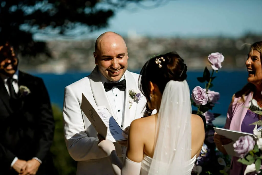 A wedding ceremony outdoors with a bride and groom exchanging vows, surrounded by bridesmaids and groomsmen, with water and city skyline in the background.