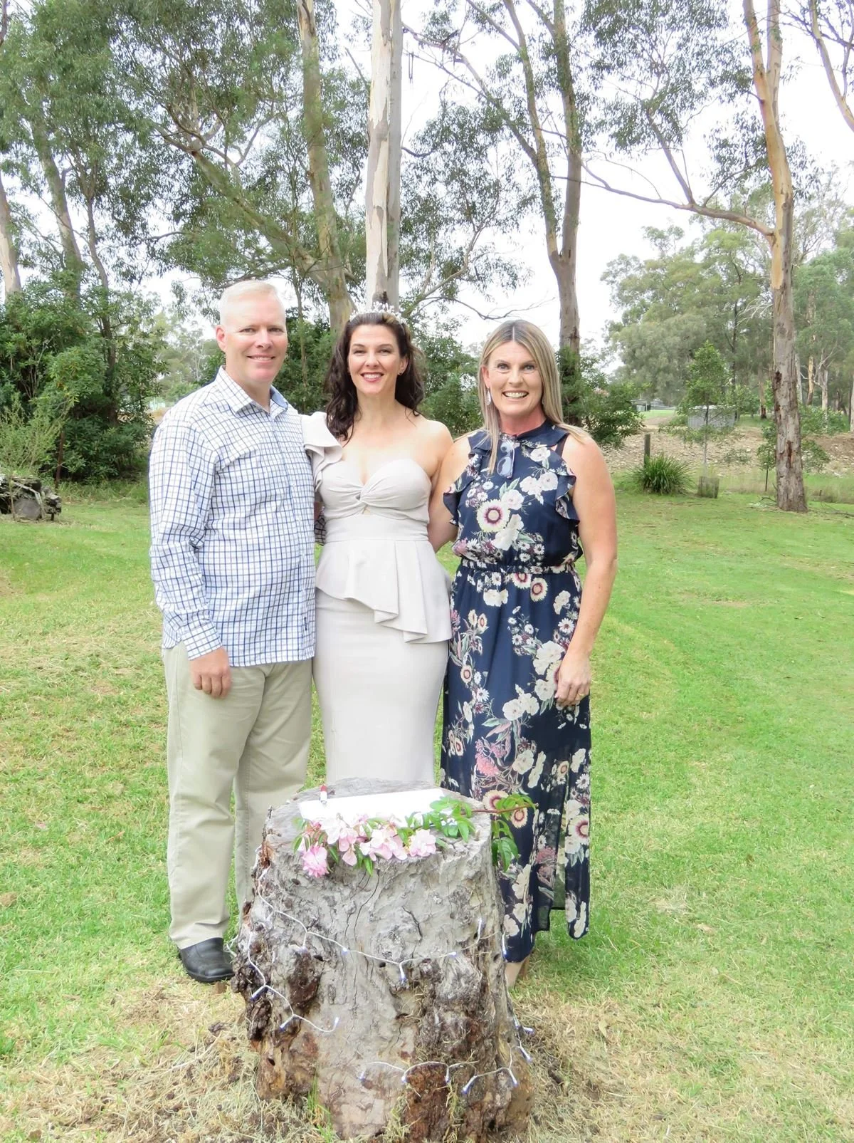 Three people standing outdoors in front of a large tree, near a rustic tree stump decorated with flowers and string lights, with a grassy field and trees in the background. One woman is wearing a white dress, and the other woman is in a floral dress, next to a man in a checkered shirt and khakis.