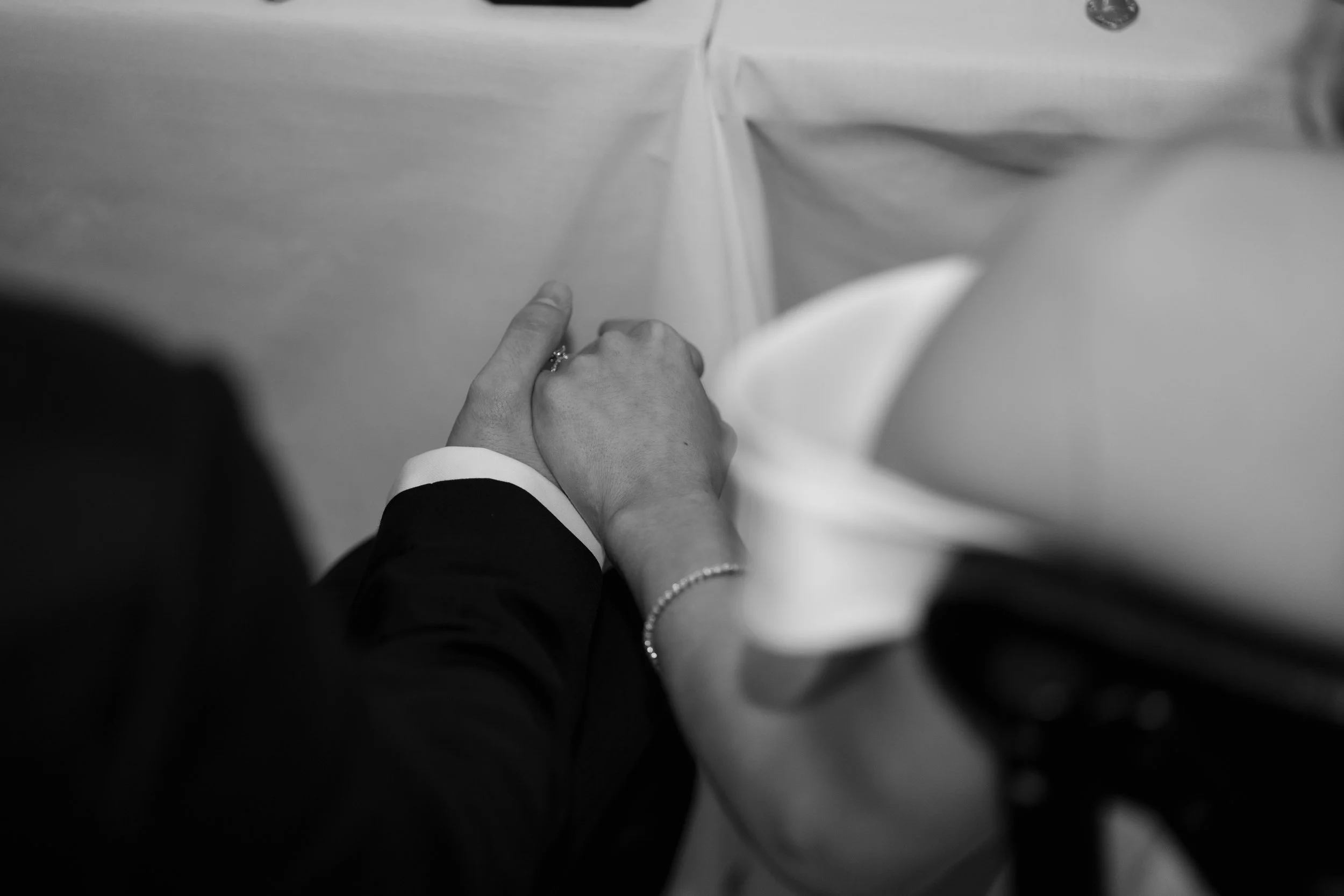 Close-up of a couple holding hands during a wedding ceremony, with the bride wearing a pearl bracelet and the groom in a suit, in black and white.
