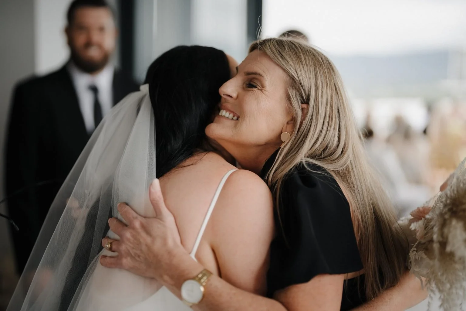 A woman in a wedding dress hugging an older woman with gray hair, who is smiling warmly. Another man in a suit is in the background, smiling.