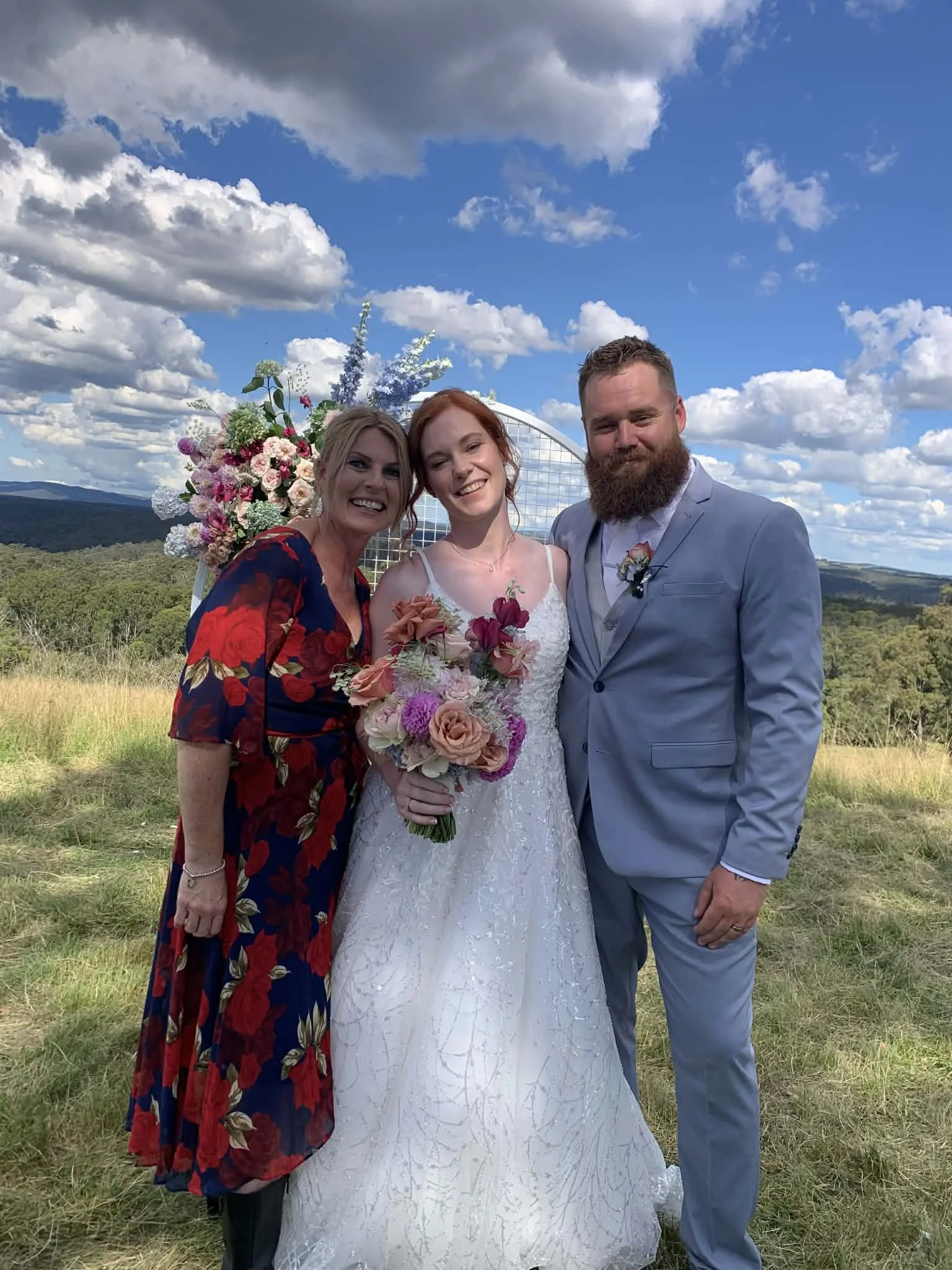A bride and groom standing outdoors on a grassy hill with a scenic landscape and blue sky with clouds in the background, accompanied by a woman in a floral dress, all smiling.