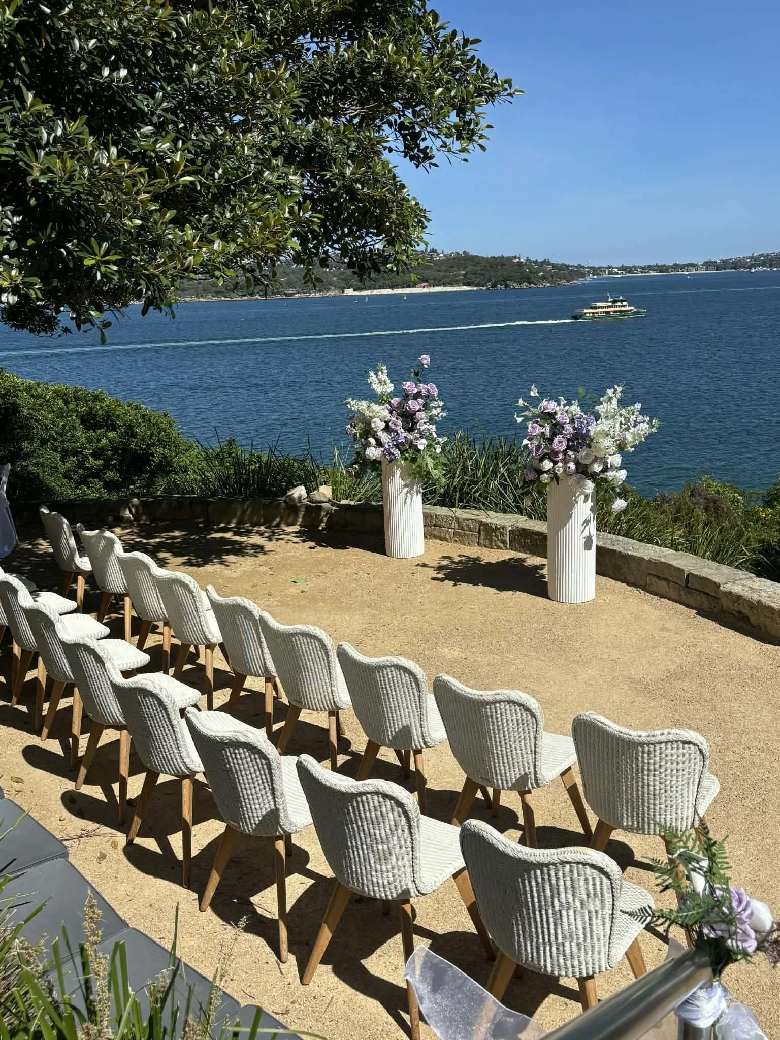 Set up for an outdoor wedding ceremony overlooking a body of water with a boat passing by, featuring white chairs and flower arrangements in vases.