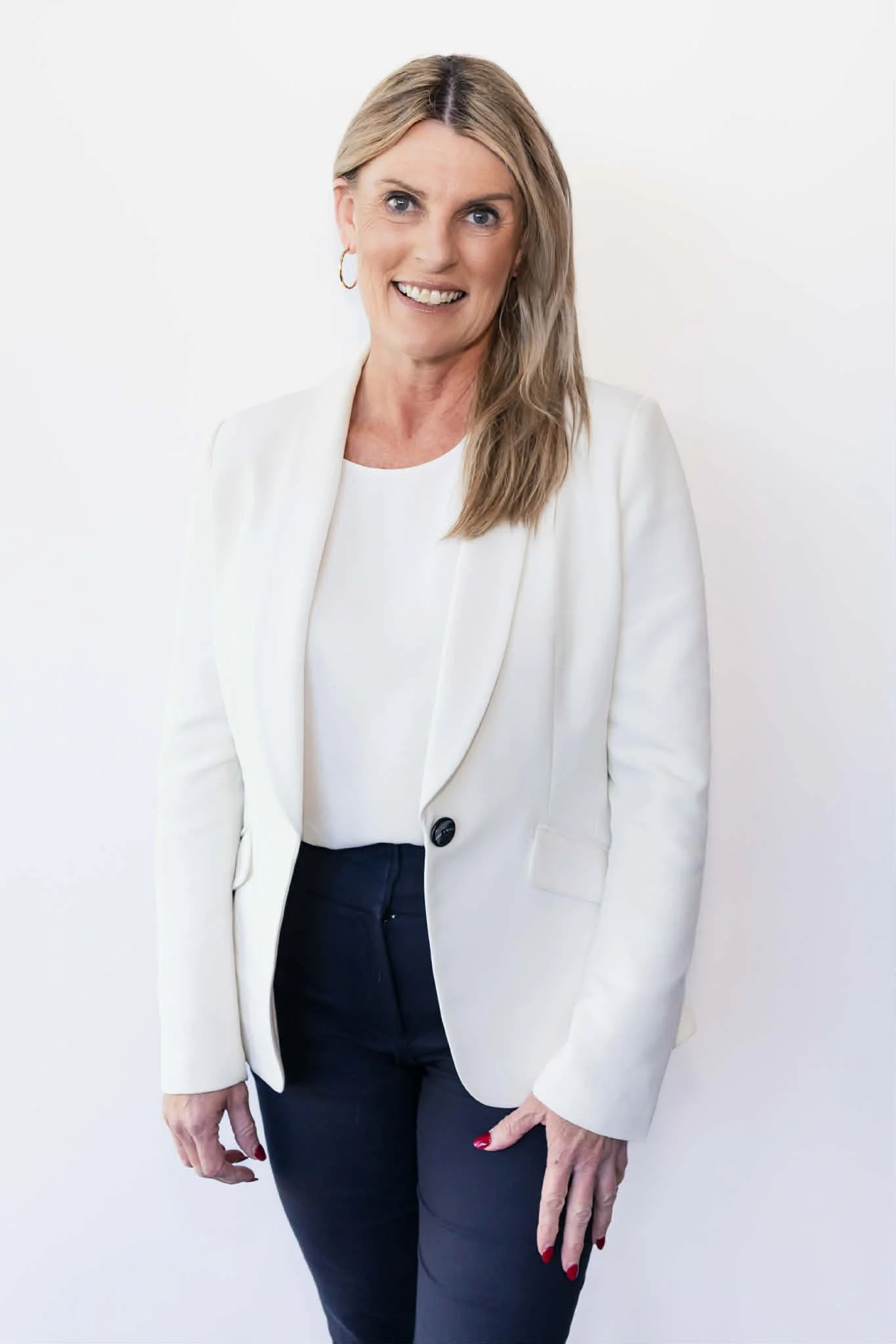 A woman with blonde hair in a white blazer and black pants, smiling, standing against a plain white background.