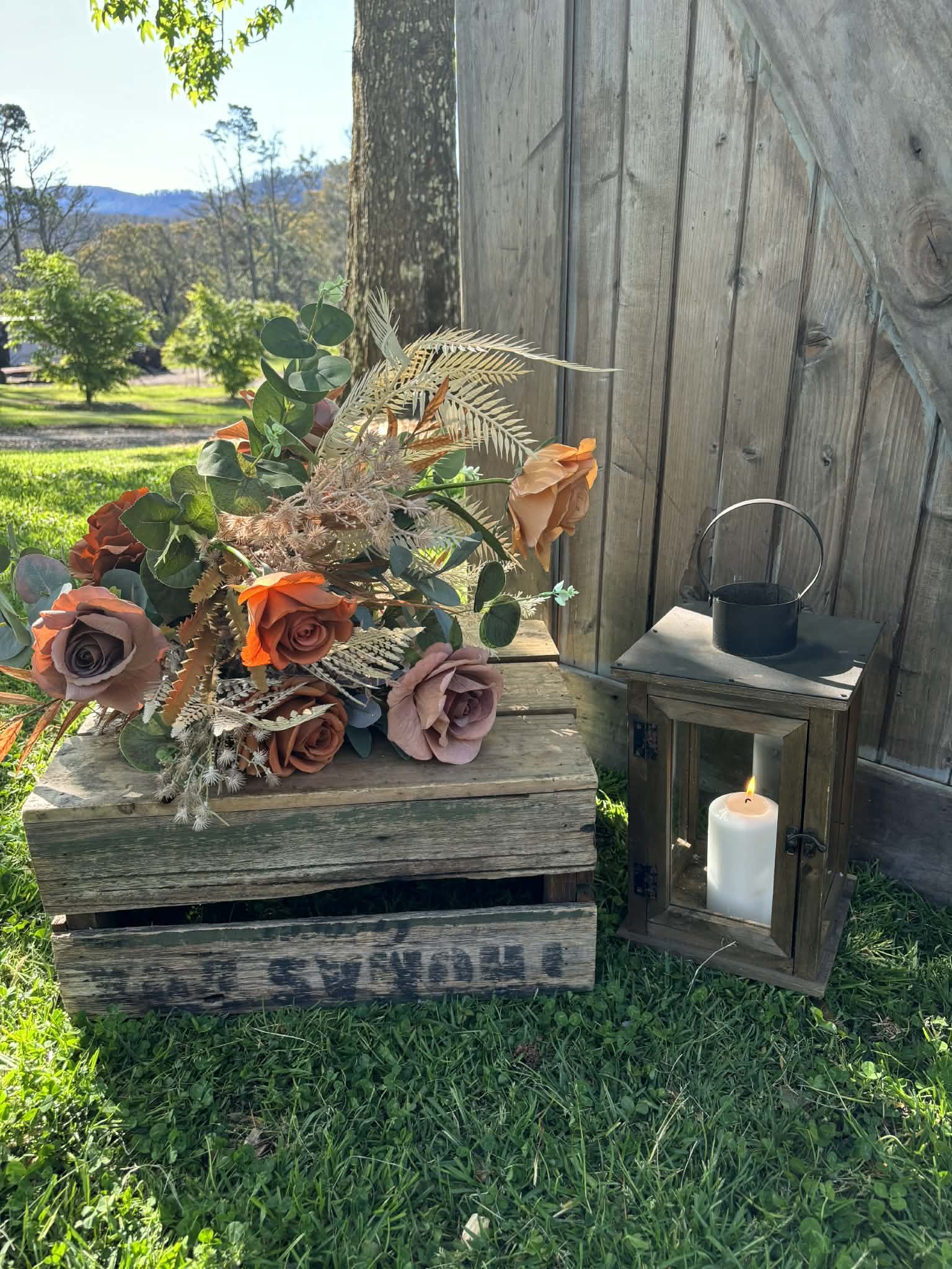 A rustic outdoor scene featuring a bouquet of artificial roses and foliage on a weathered wooden crate, next to a wooden lantern with a burning white candle inside, and a small black bucket on a table, set against a wooden fence with a grassy area and trees in the background.