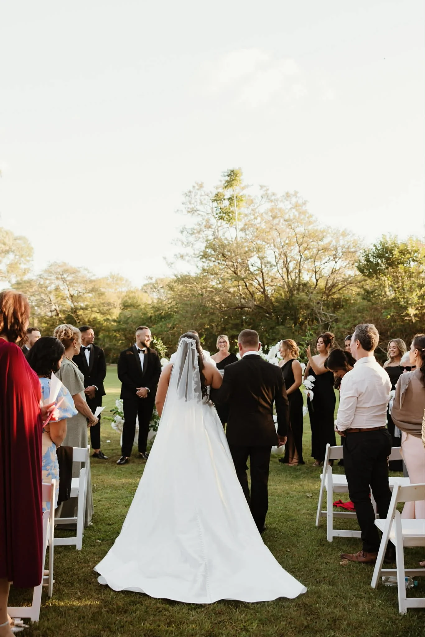 Bride and groom walking down the aisle at an outdoor wedding ceremony, with guests standing on either side, during the daytime with clear skies and trees in the background.