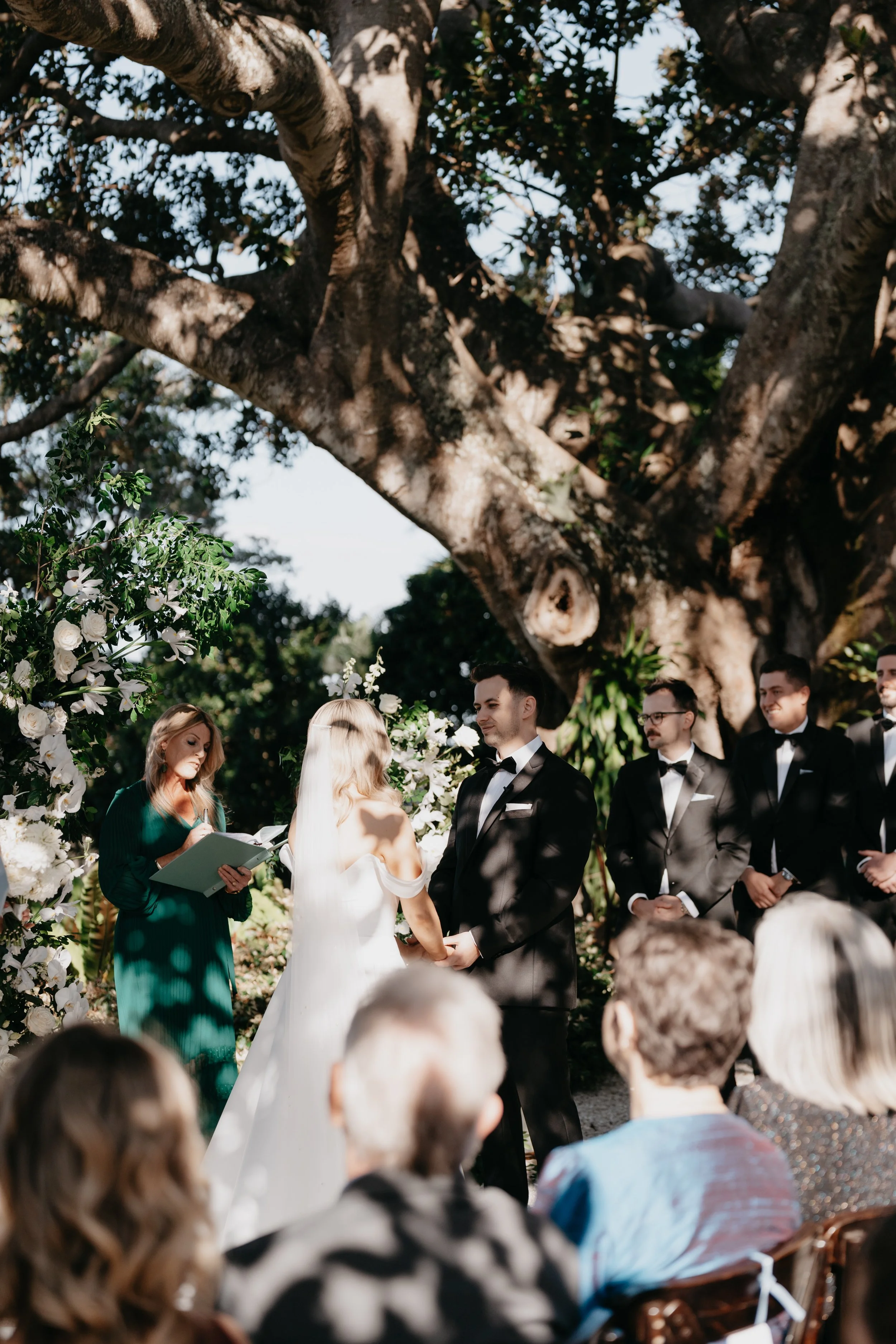 A wedding ceremony outdoors under a large tree, with the bride and groom holding hands, exchanging vows, and officiant reading from a book, surrounded by wedding party and guests.
