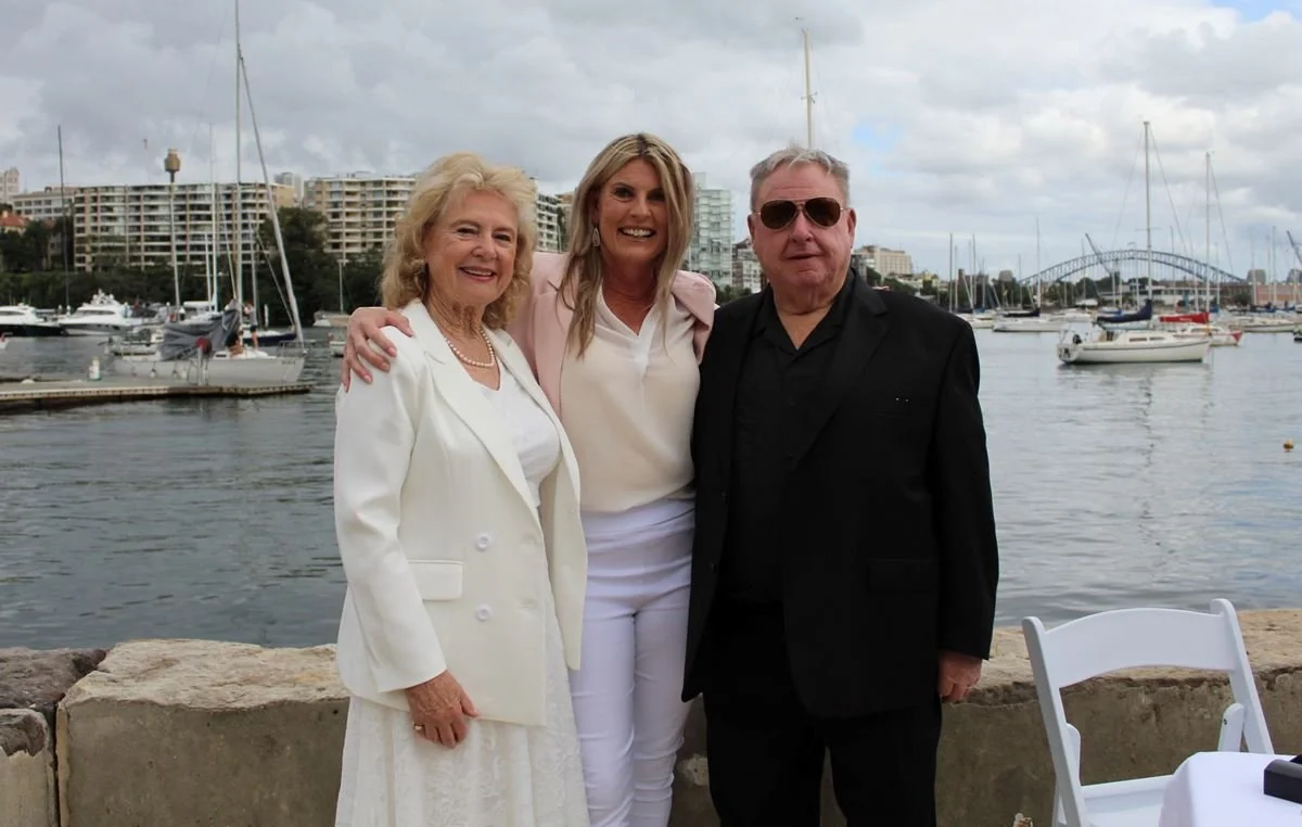 Three adults, two women and one man, standing together outdoors near a waterfront with boats, high-rise buildings, and a bridge in the background.