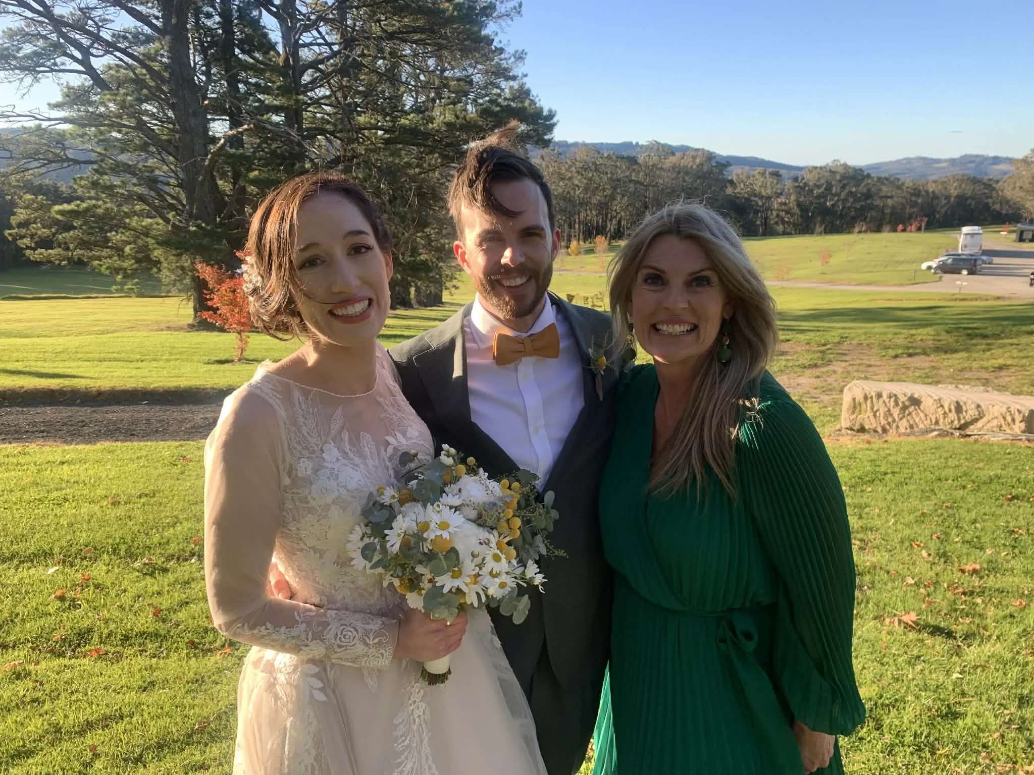 Three people smiling outdoors during a wedding celebration, with a woman in a wedding dress holding a bouquet, a man in a suit and bow tie, and another woman in a green dress, standing on a grassy area with trees and hills in the background.