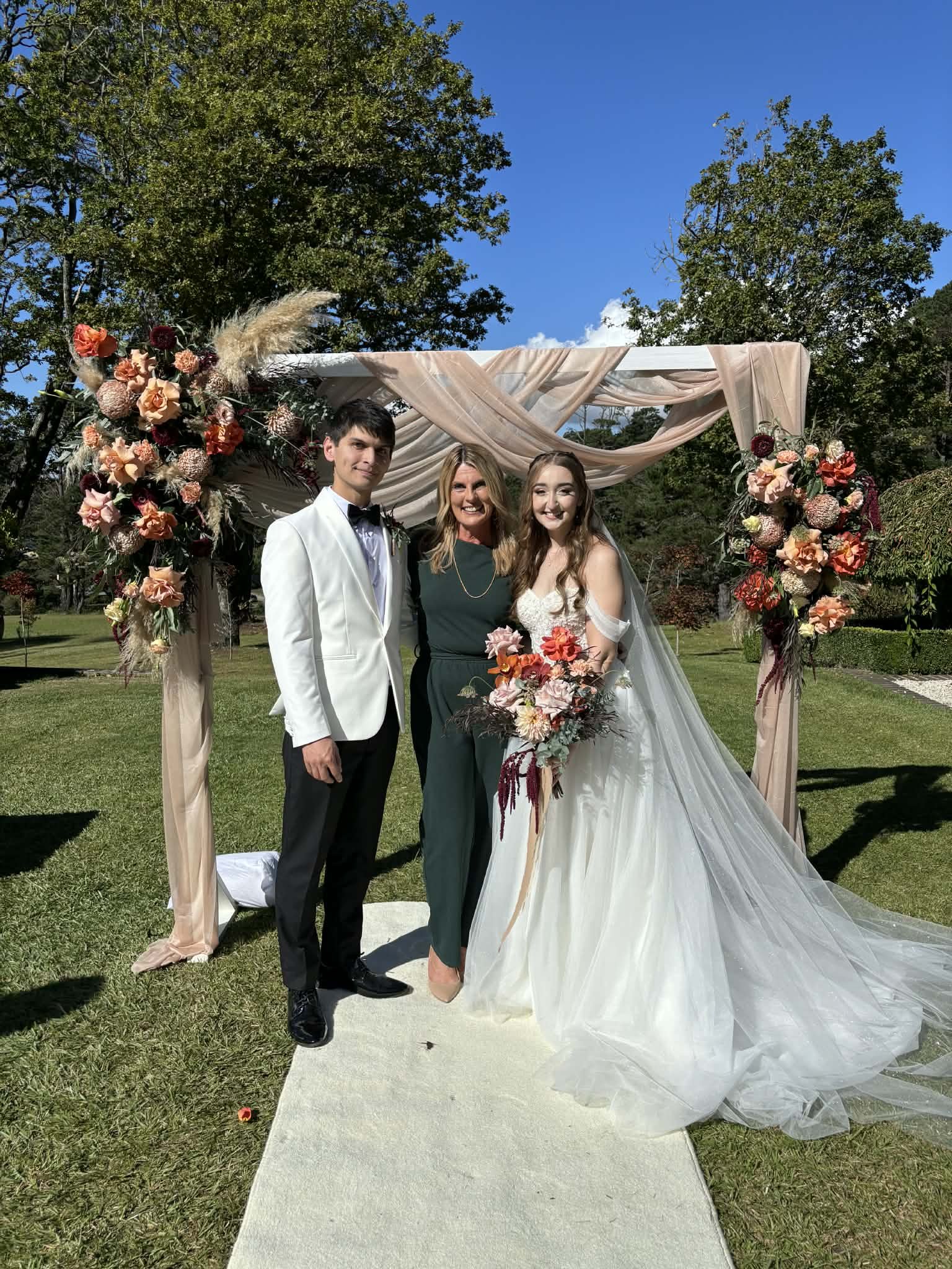 A bride and groom with a woman, possibly a wedding officiant or family member, standing under a floral wedding arch outdoor on a sunny day.