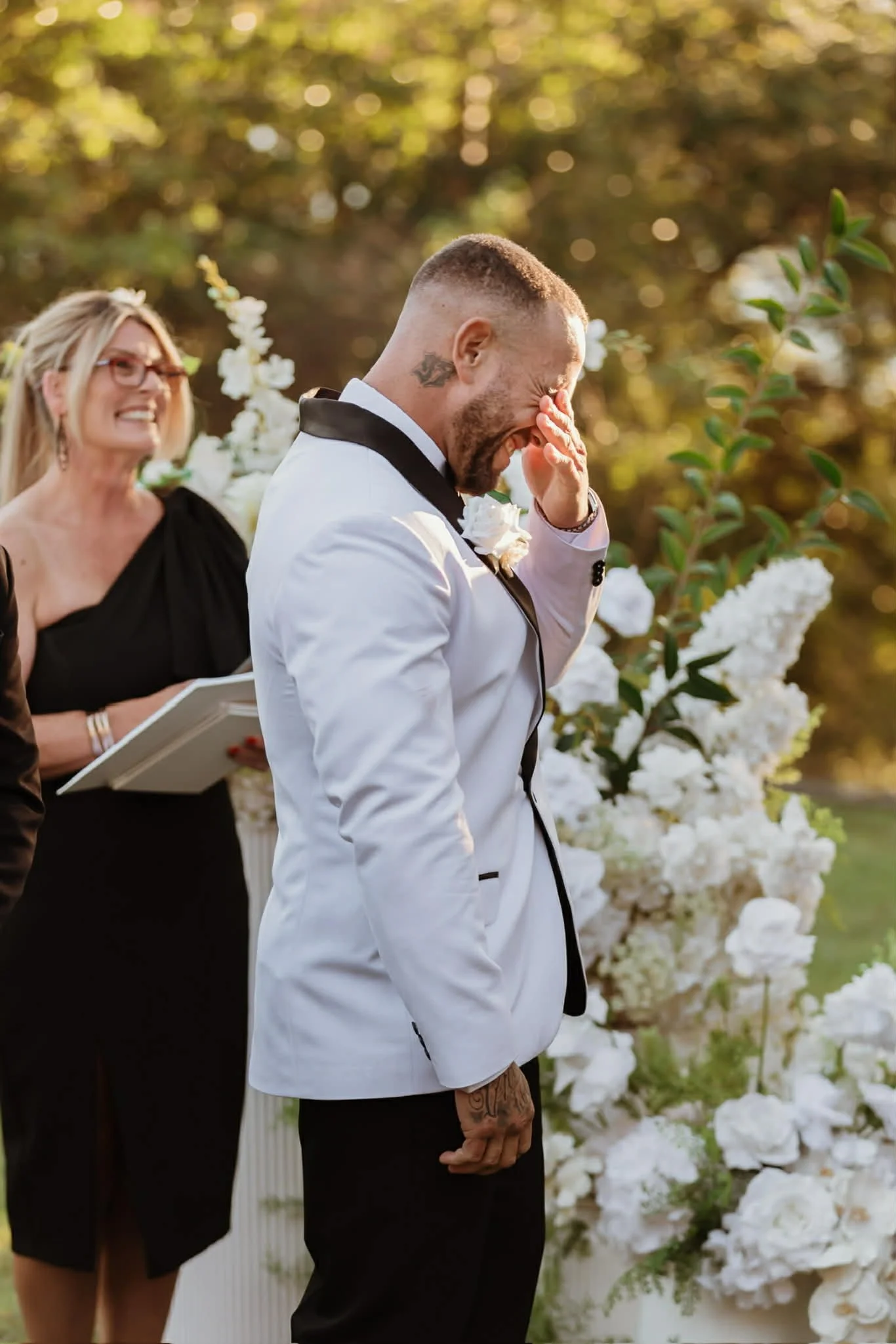 A man in a white tuxedo with black accents, covering his face and from a joyous tear, stands outdoors during a wedding ceremony with a woman officiating and white floral decorations around him.