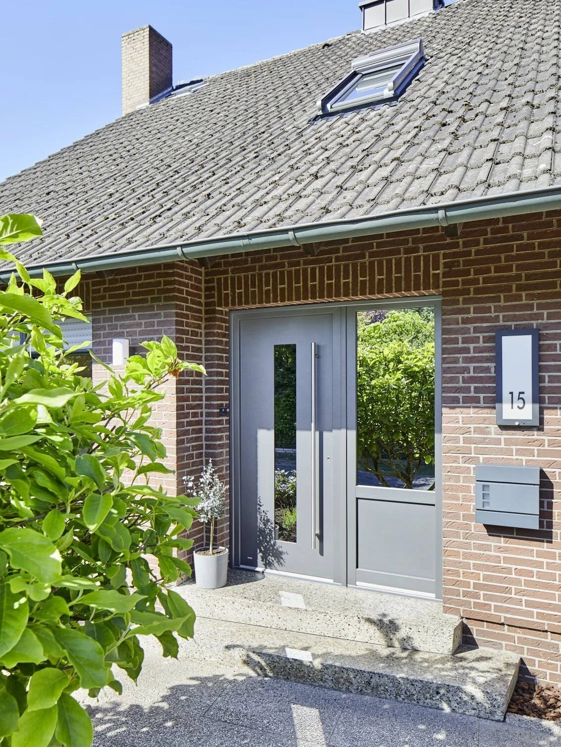 Moderner Eingang zu einem Haus mit grauser Tür, großen Fenstern, Briefkasten und Hausnummer 15, umgeben von grünen Pflanzen und einem Ziegelmauer.