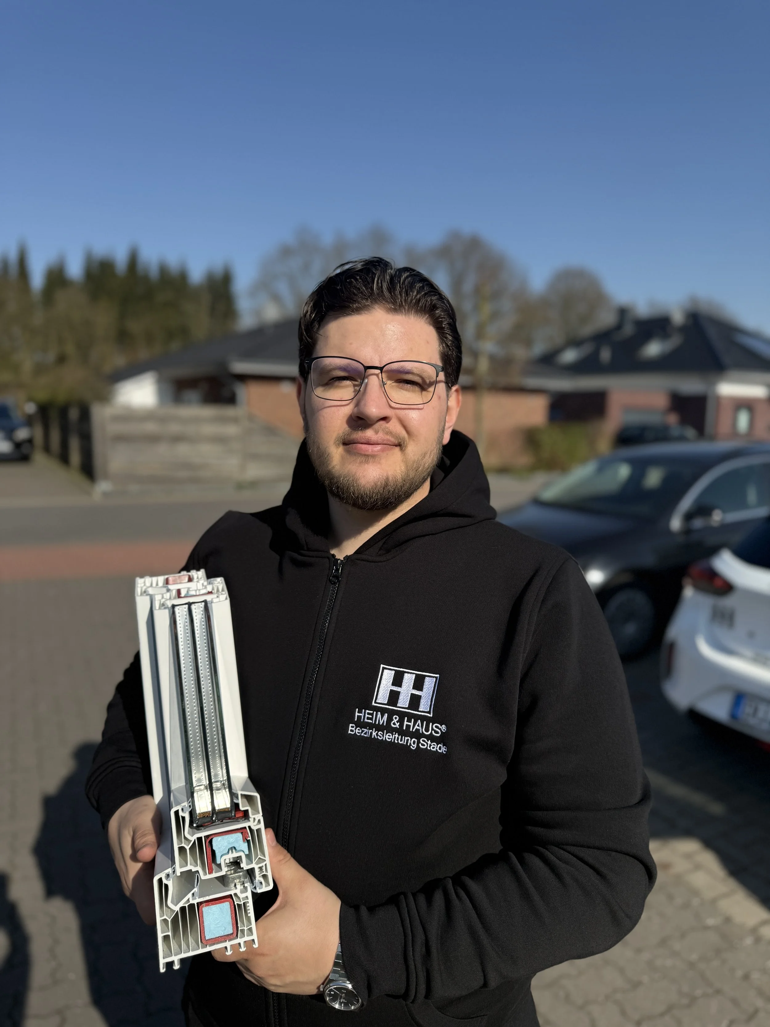 Junger Mann trägt eine schwarze Jacke mit dem Logo 'HEIM & HAUS' und hält eine große Kunststoff- oder Metallprofil. Er steht vor einem Parkplatz auf einer Straße bei sonnigem Wetter.