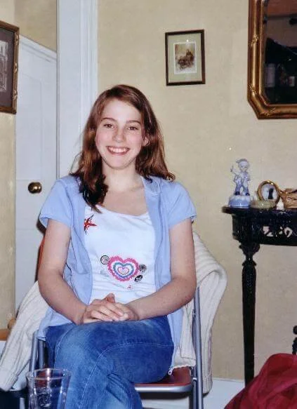 Sophie Dean Decluttering Expert and Professional Organiser when she was younger sitting on a chair in a living room, smiling at the camera, with a cream-colored wall, a framed picture, and a mirror in the background.