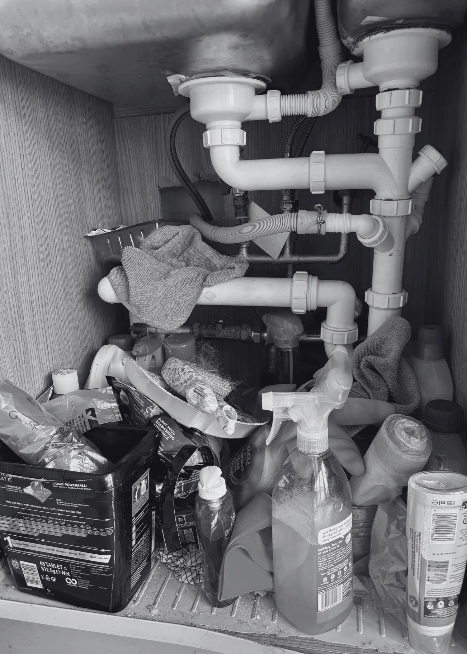 A messy kitchen cupboard under the sink with cleaning products, cloths and dishwasher tablets