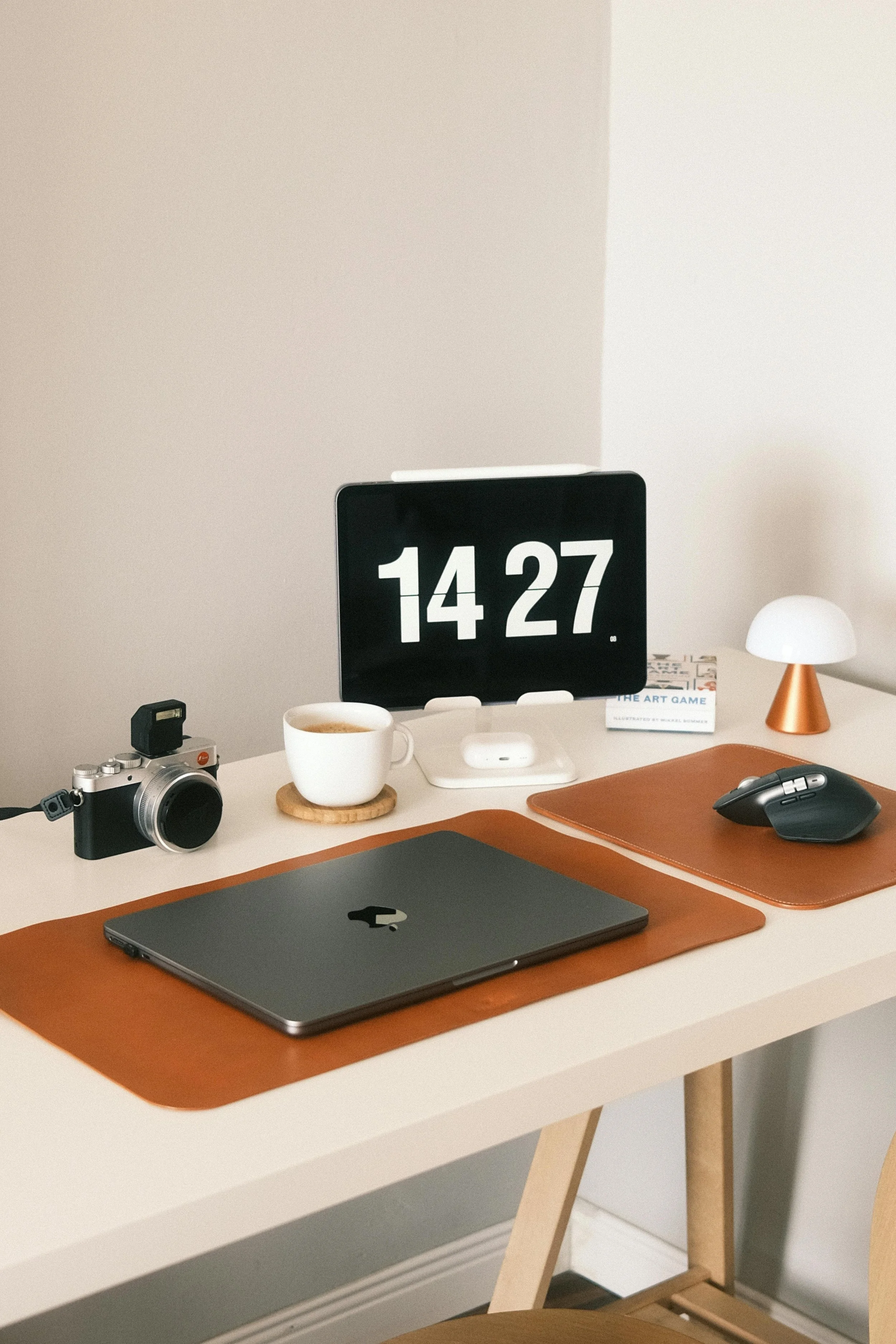 A minimalist desk with two brown desk pads with a laptop and a mouse on each. A tablet at the back displays the time, the desk also has a camera, coffee mug and small lamp on it.