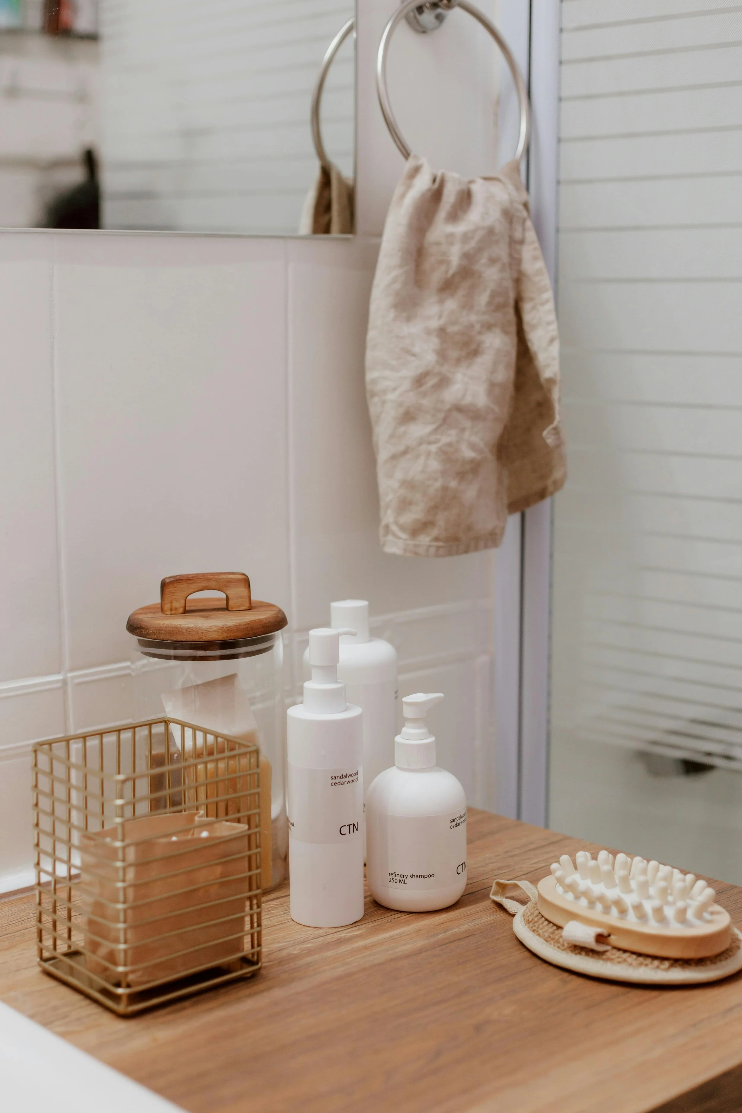 Bathroom countertop with skincare bottles, a gold wire basket with cotton pads, a wooden hairbrush, and beige towels hanging on ring hooks behind a mirror.