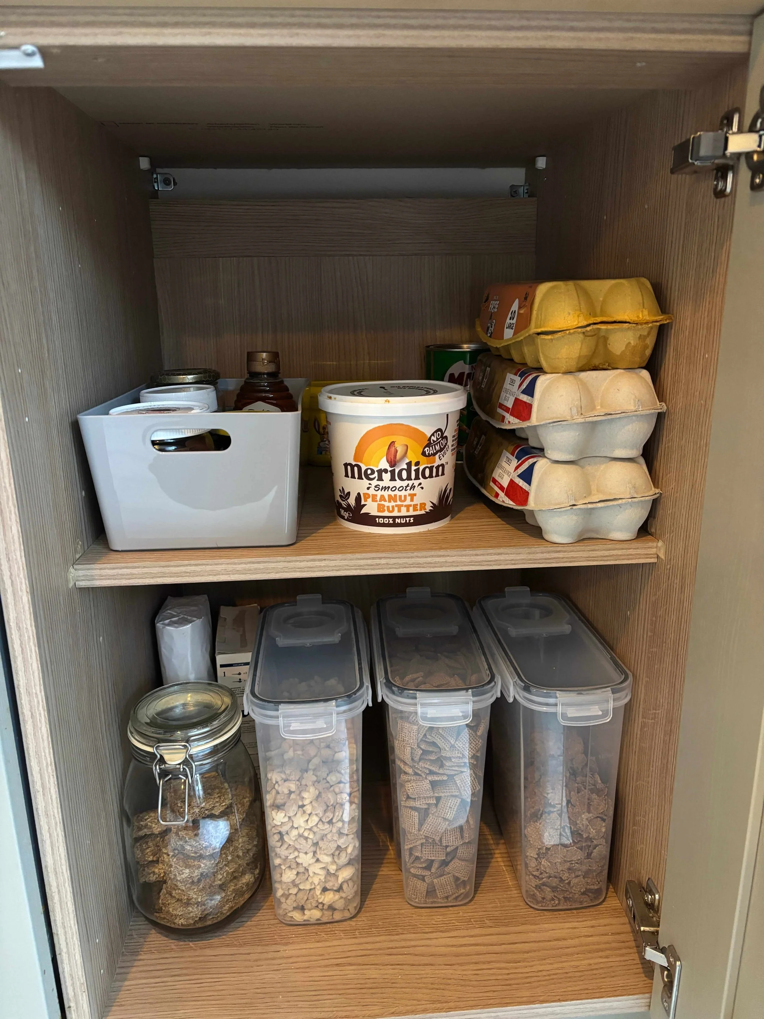 An organised kitchen cupboard with breakfast cereals in containers, spreads and honeys in a container and eggs in cartons.