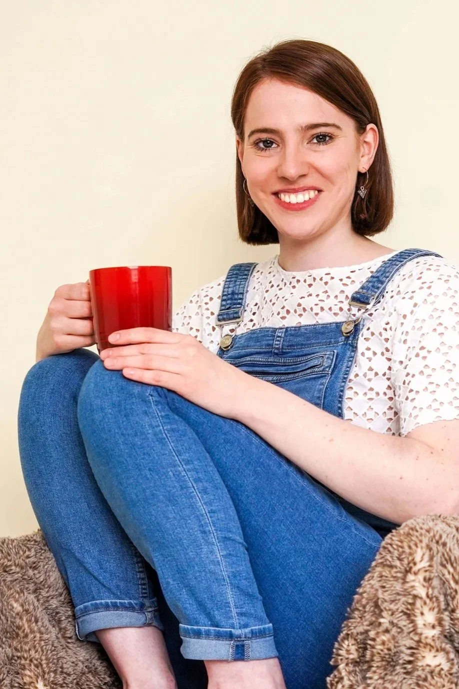 A young woman with short brown hair, smiling, wearing a white lace top and blue overalls, sitting on a blanket and holding a red mug.