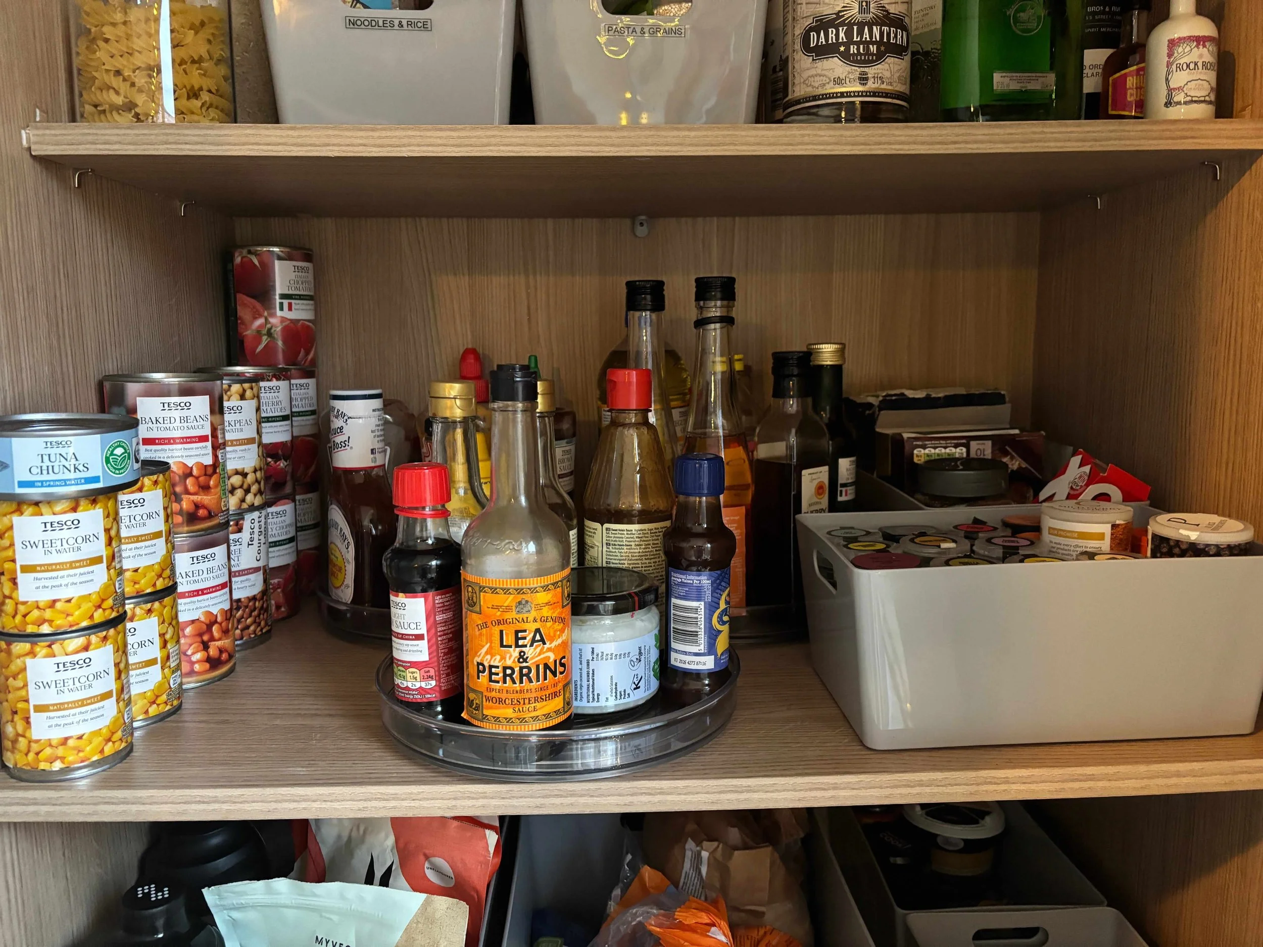 Pantry shelf with canned foods, condiments, bottles of sauces and vinegar, and a white container filled with coffee pods.