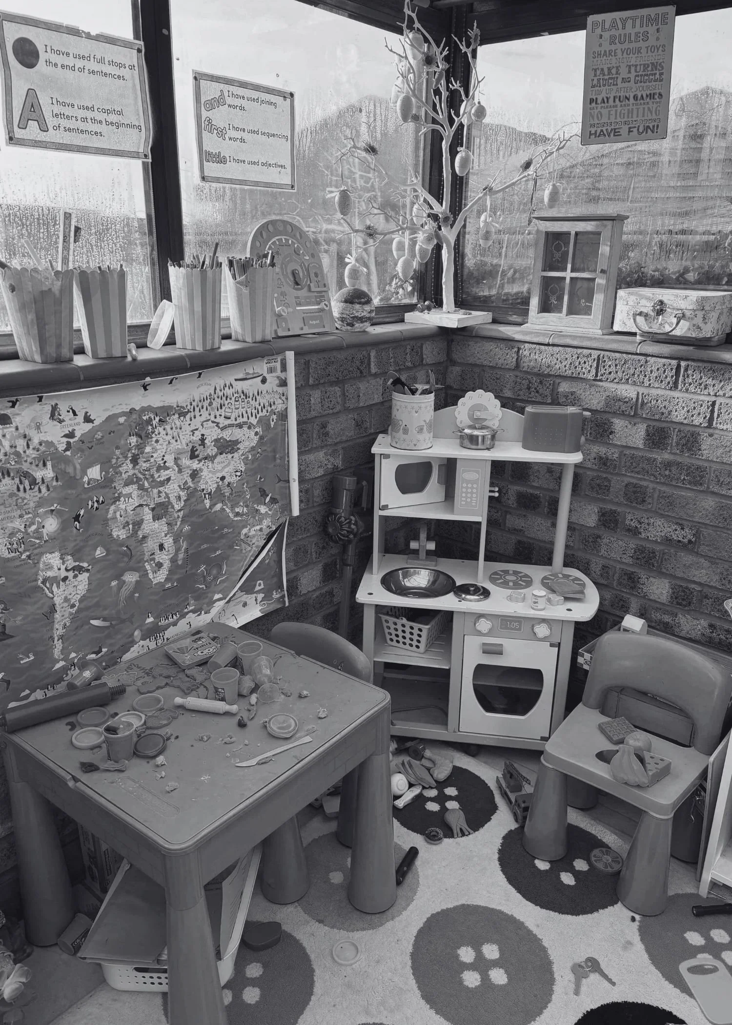 A messy corner in a children's playroom with a play kitchen with wooden food on it, a table with playdough on it and chairs around it.