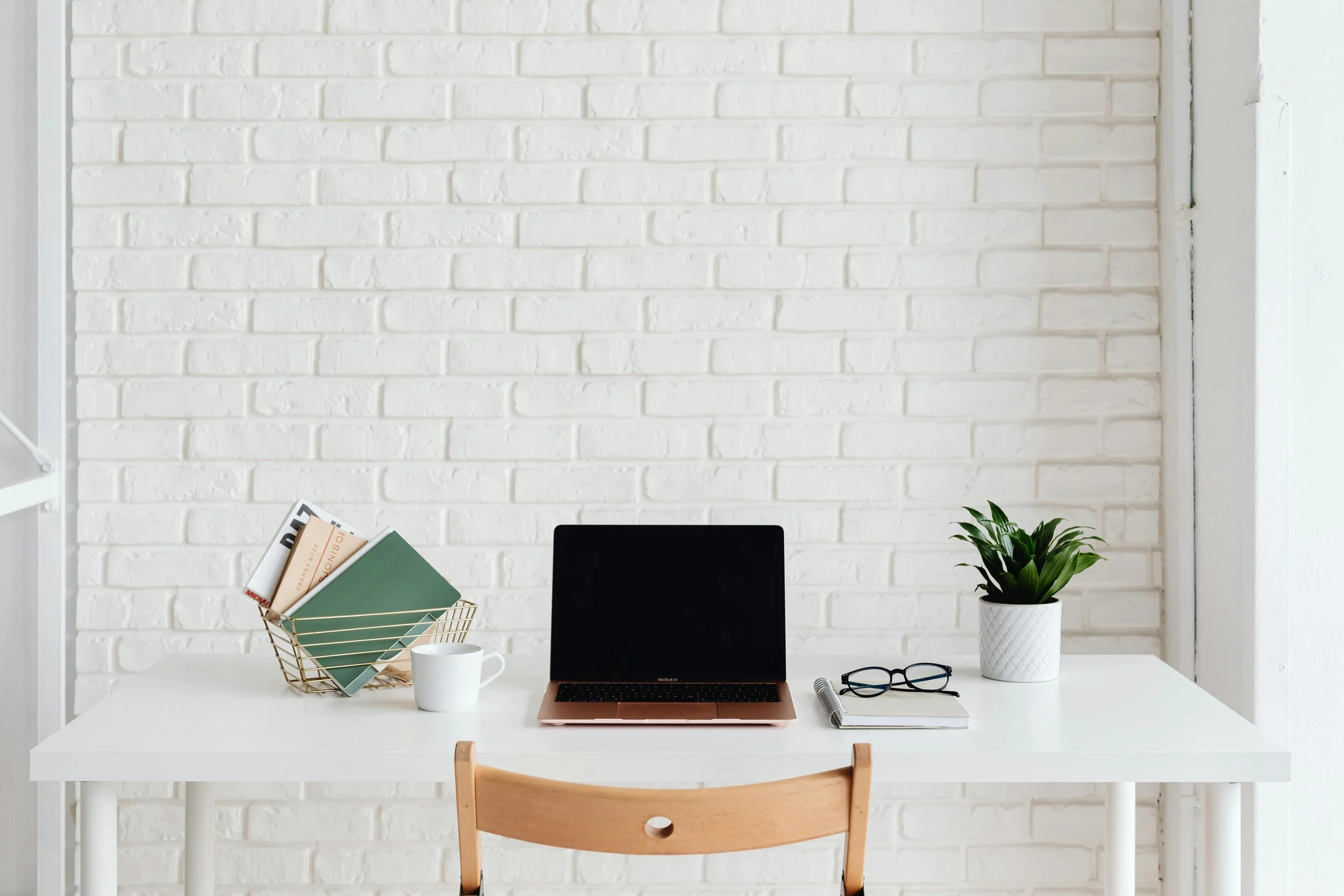 Minimalist white desk against a white brick wall with a laptop, a pair of glasses on a notebook, a potted plant, a white coffee mug, and a wire basket with magazines.