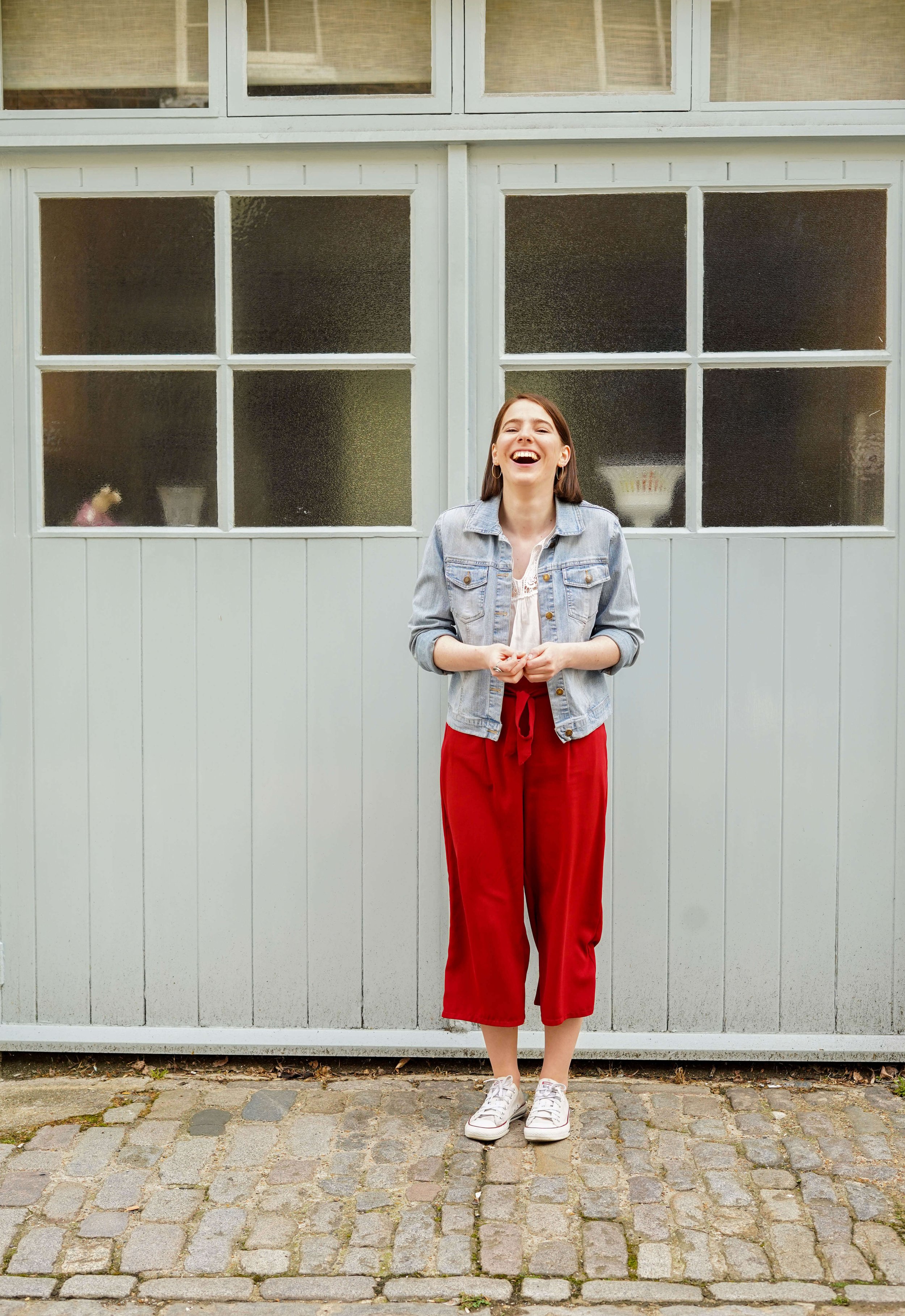 A young woman with shoulder-length brown hair, wearing a light denim jacket, white top, red pants, and white sneakers, standing and laughing in front of a light gray wooden wall with large windows.