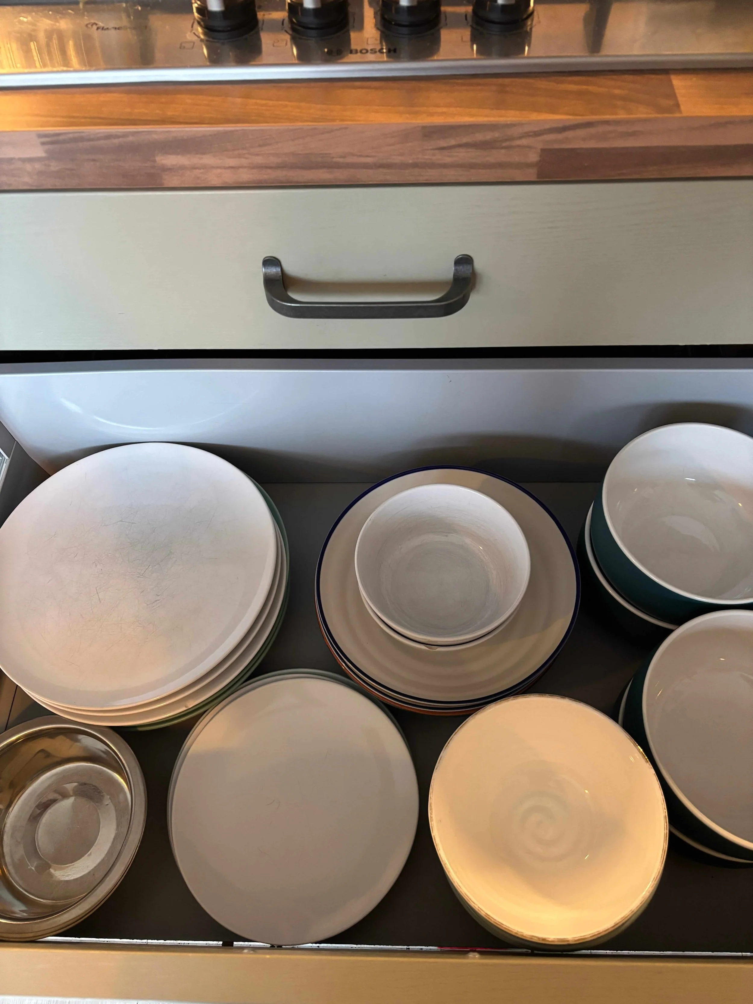 A collection of stacked empty plates, bowls, and cups on a kitchen countertop, with a drawer and cooktop visible in the background.
