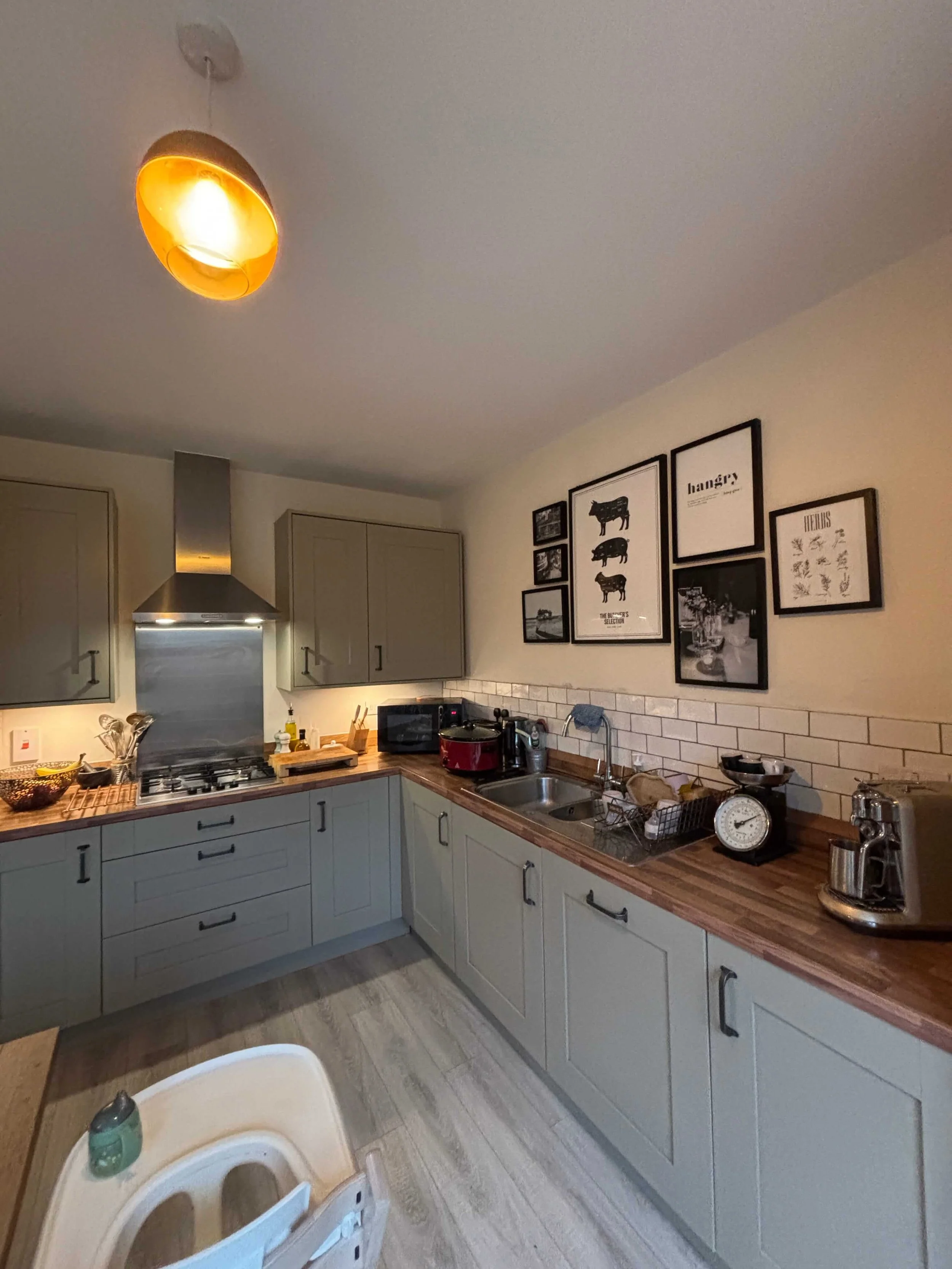 A modern kitchen with gray cabinets, a wooden countertop, a stainless steel range hood, and white subway tile backsplash. Decor includes black-framed art on the wall, various kitchen appliances and utensils on the counter, and a ceiling light with a warm glow.