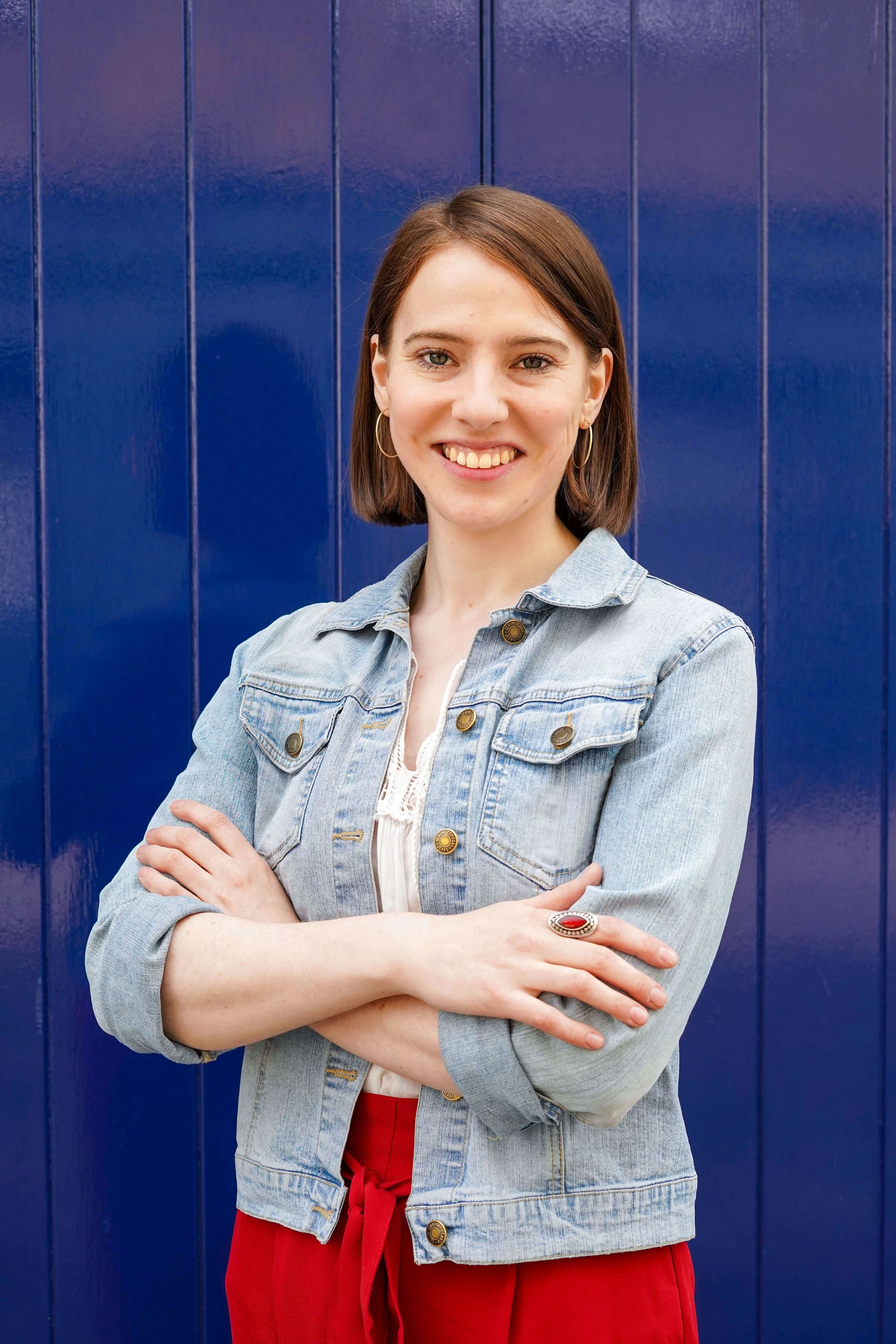 Sophie Dean Decluttering Expert and Professional Organiser with short brown hair smiling standing in front of a blue wall with vertical panels, wearing a light denim jacket, a white top, and red trousers.