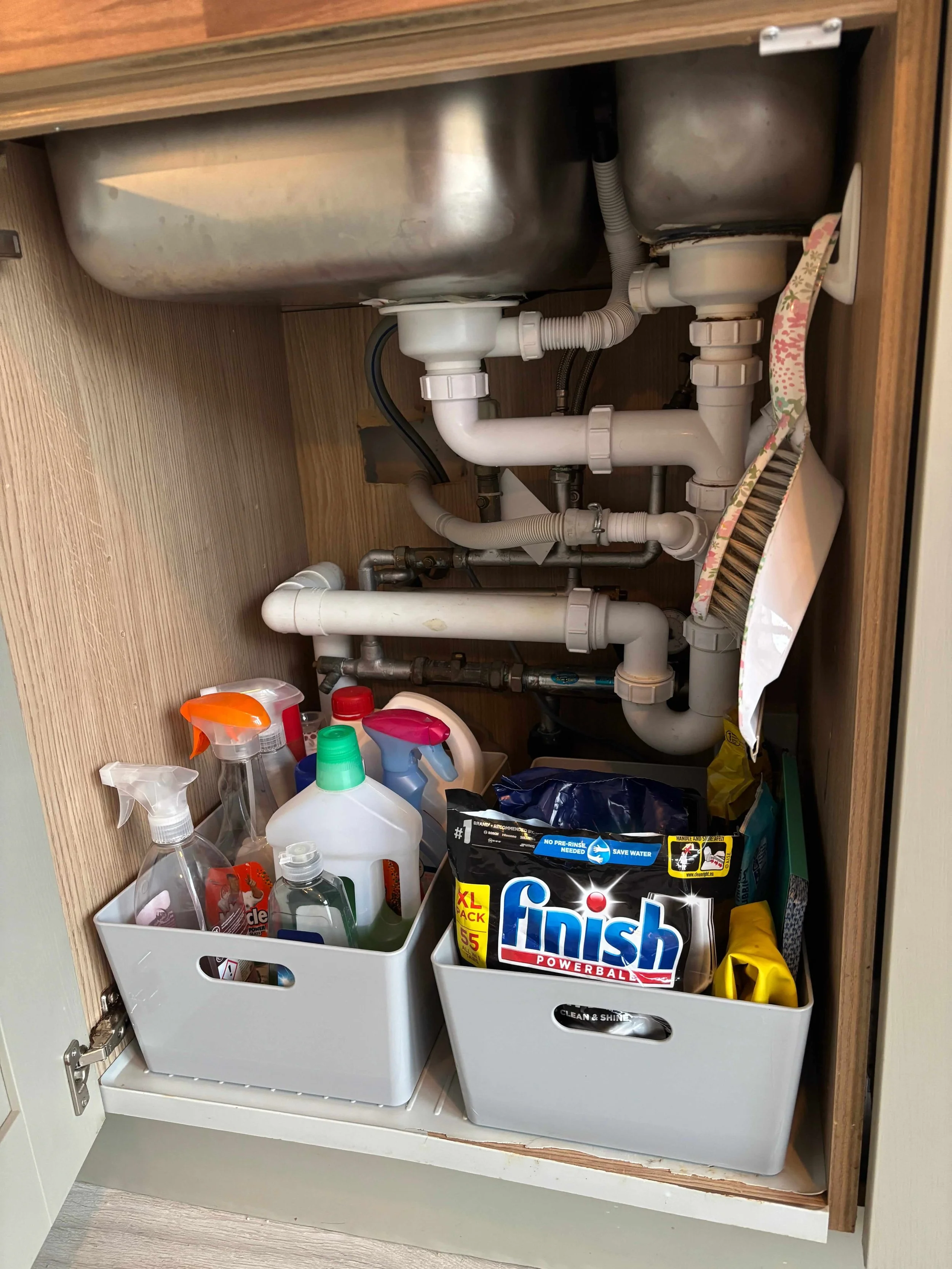 An organised under the sink cupboard in a kitchen with cleaning sprays, cloths and dishwasher tablets in plastic boxes and a dustpan and brush on a hook.