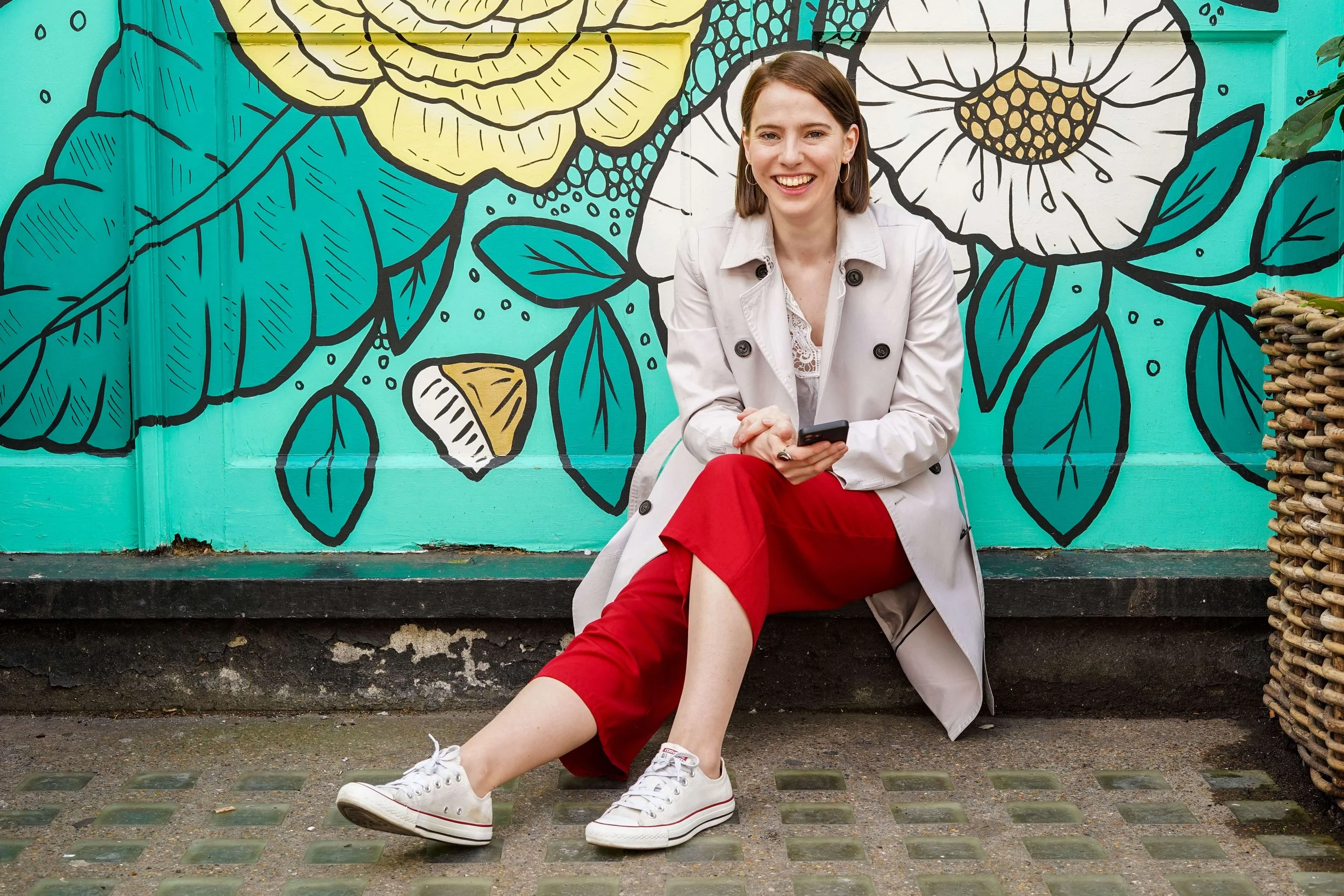 Sophie Dean Decluttering Expert and Professional Organiser with shoulder length brown hair sitting on the ground smiling while holding a smartphone against a colourful mural with large floral designs and leaves.