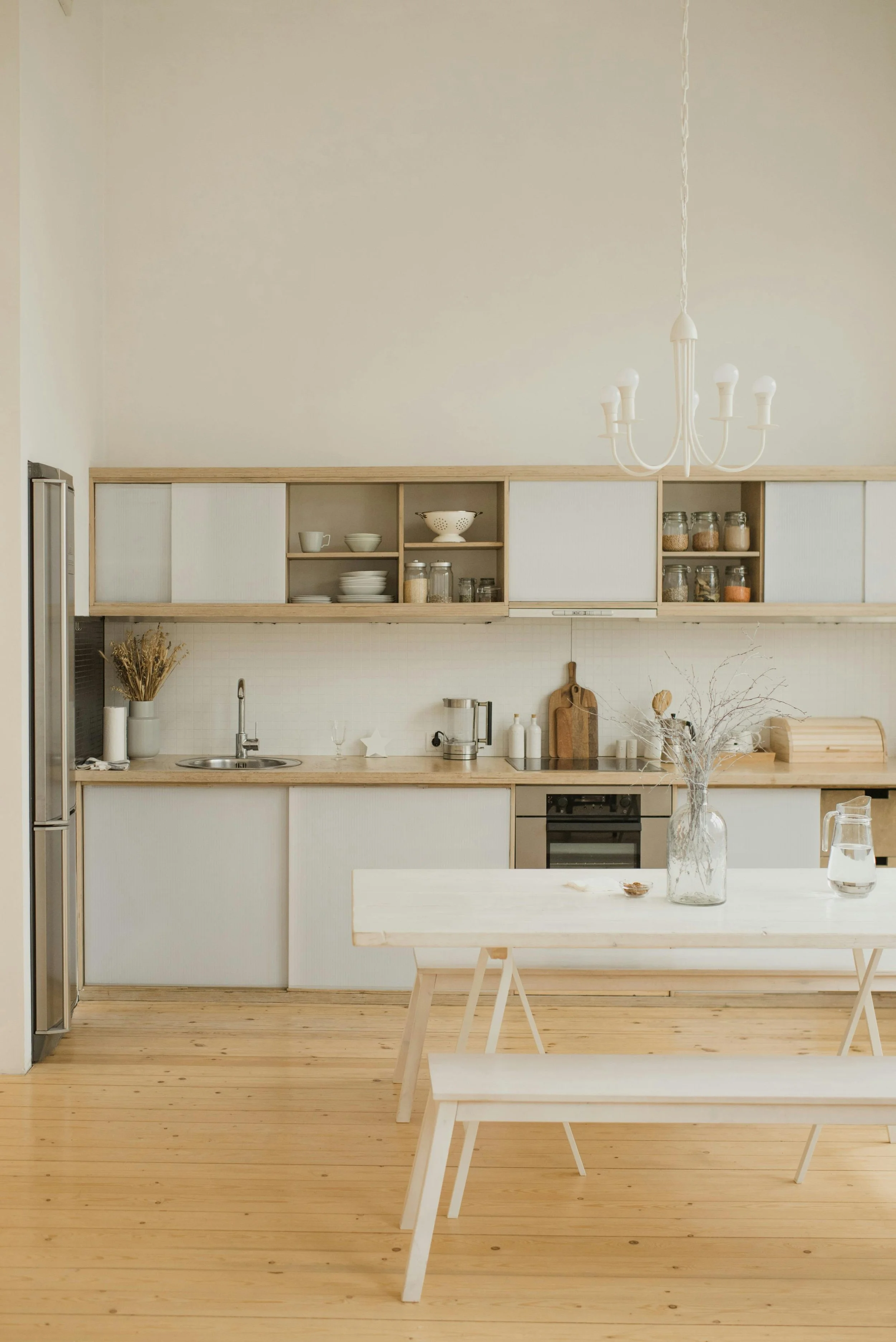 A minimalist kitchen with light wood cabinets, open shelves with dishes and jars, a wooden dining table with a bench, a glass vase with dried branches, and a white chandelier.