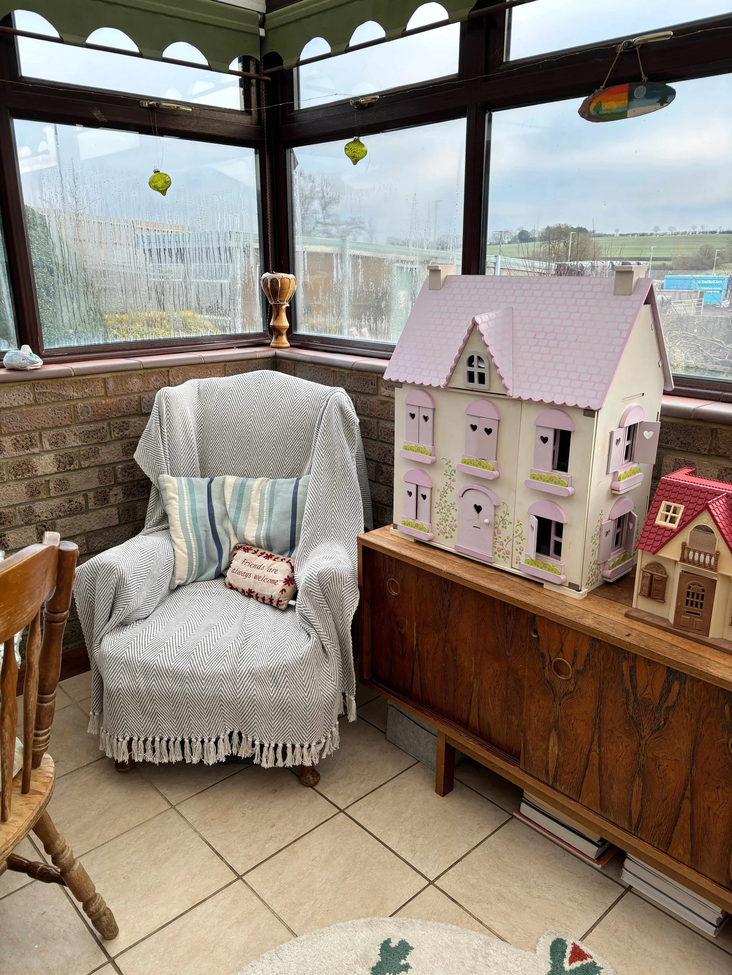 A cozy sunroom with a brick half-wall, large windowpanes, and a beige armchair with a striped pillow. On a wooden cabinet next to the armchair, there is a large decorative dollhouse with pastel colors and heart-shaped cutouts, and a smaller dollhouse with a red roof. The room has tiled flooring and some holiday decorations.