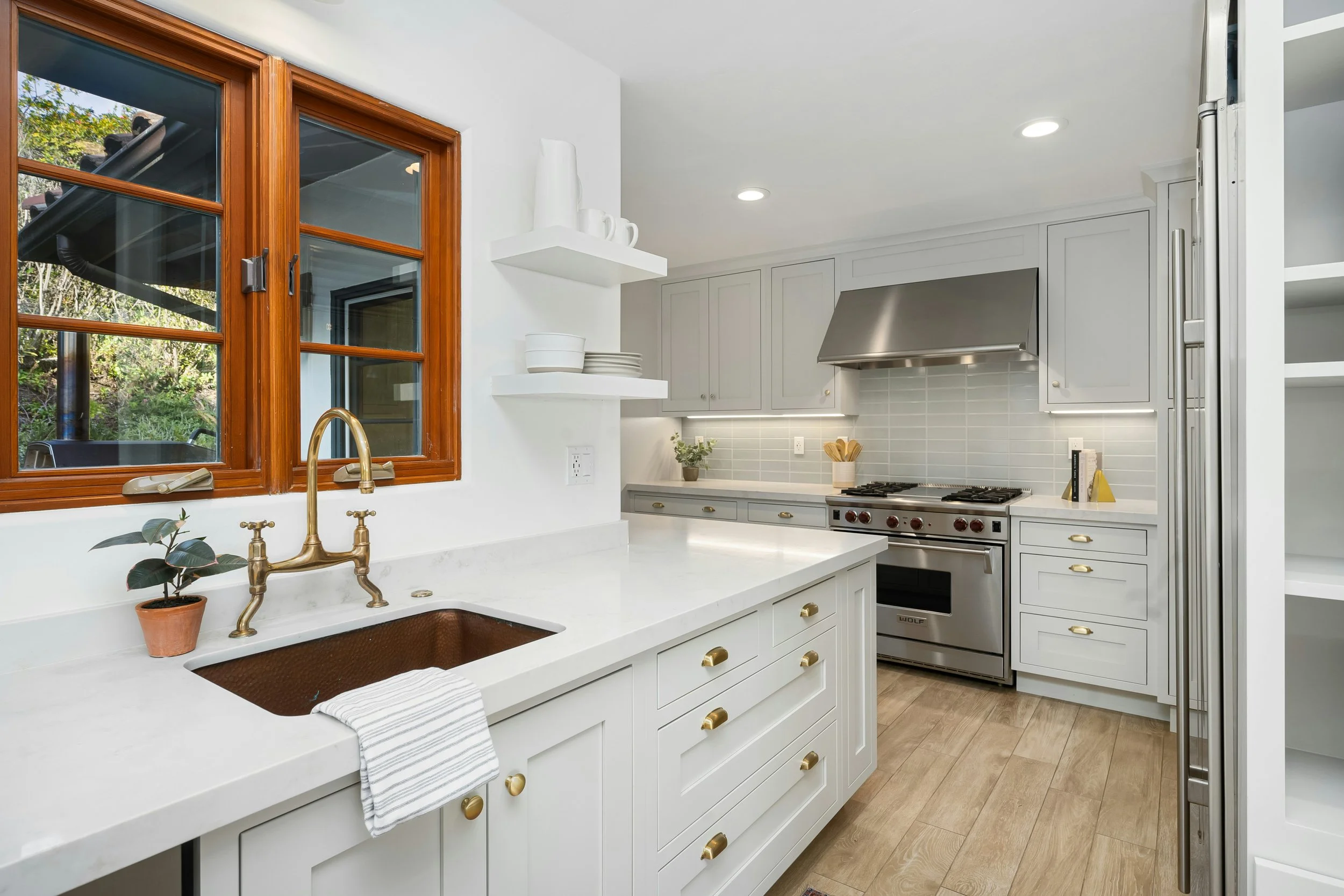 Modern kitchen with white cabinets, wooden floor, and a window showing greenery outside. Features include a brass faucet, a copper sink, open shelves with white dishes and vases, a stove with stainless steel range hood, and built-in pull-out pantry shelves.