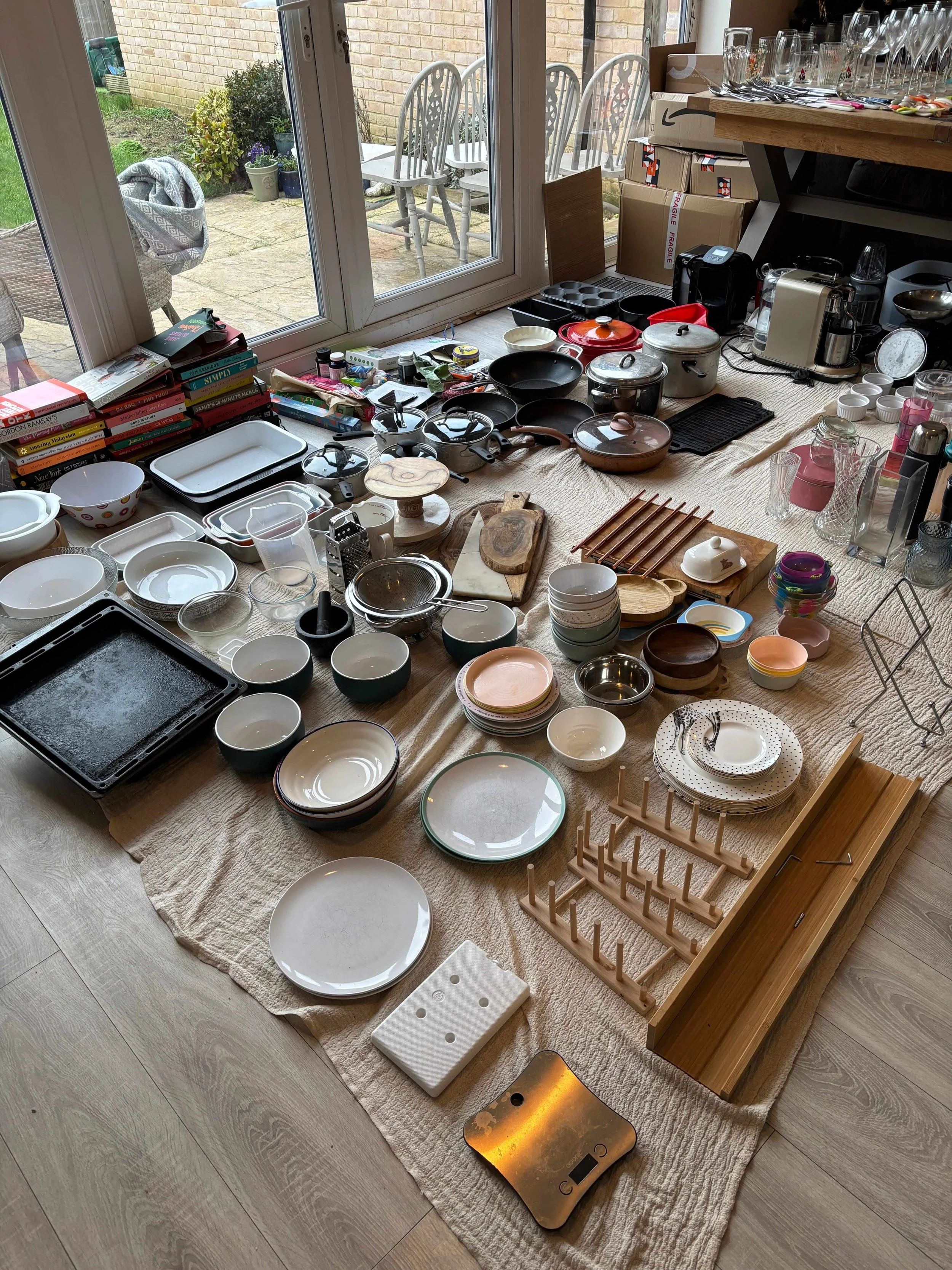 A variety of kitchenware, including plates, bowls, pots, pans, and utensils, is spread out on a large fabric covering the floor inside a room with a glass door leading to an outdoor patio.