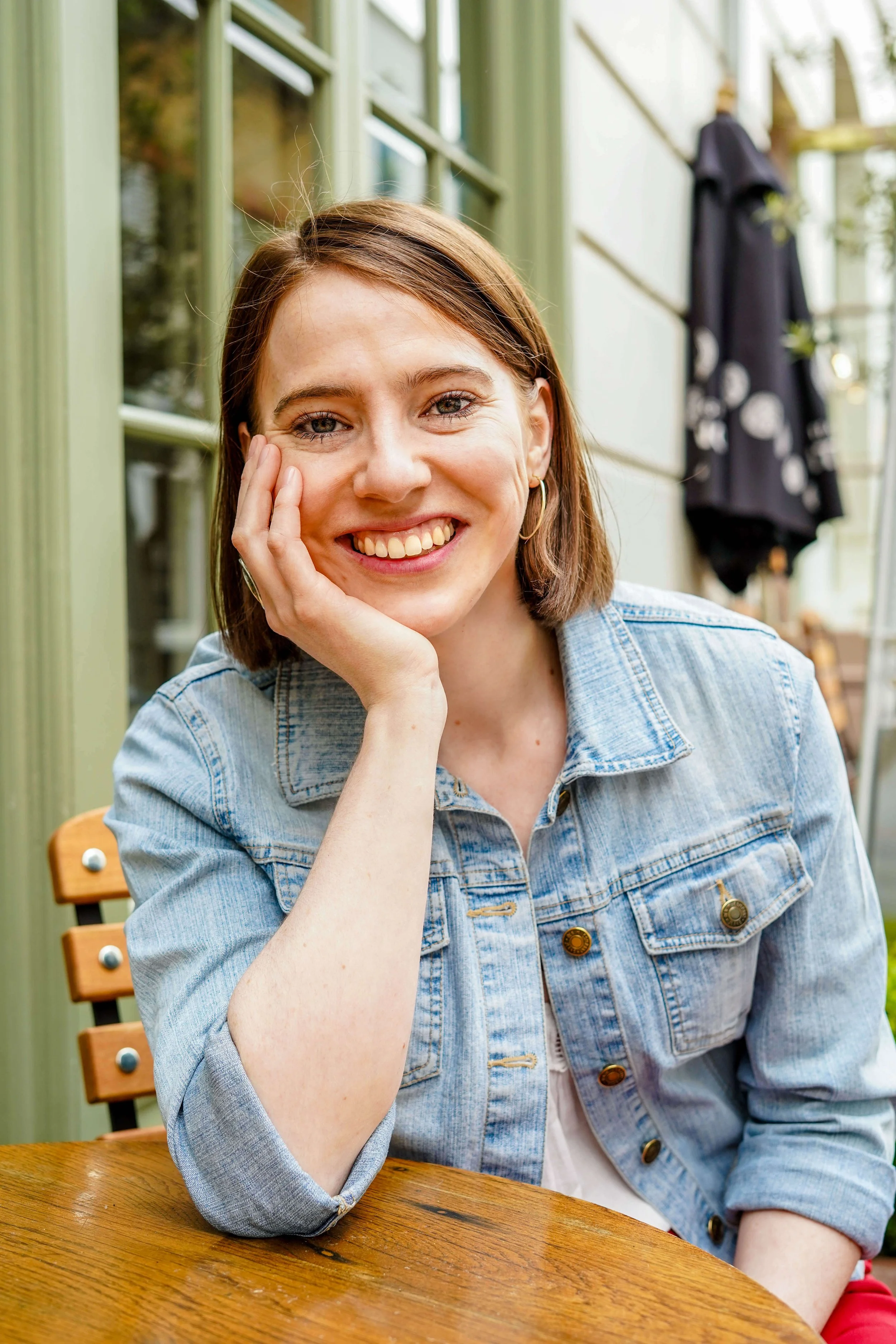 Sophie Dean Decluttering Expert and Professional Organiser with shoulder-length brown hair, smiling, sitting at a wooden table outdoors, resting her face on her hand, in front of a building with green trim and a large window.