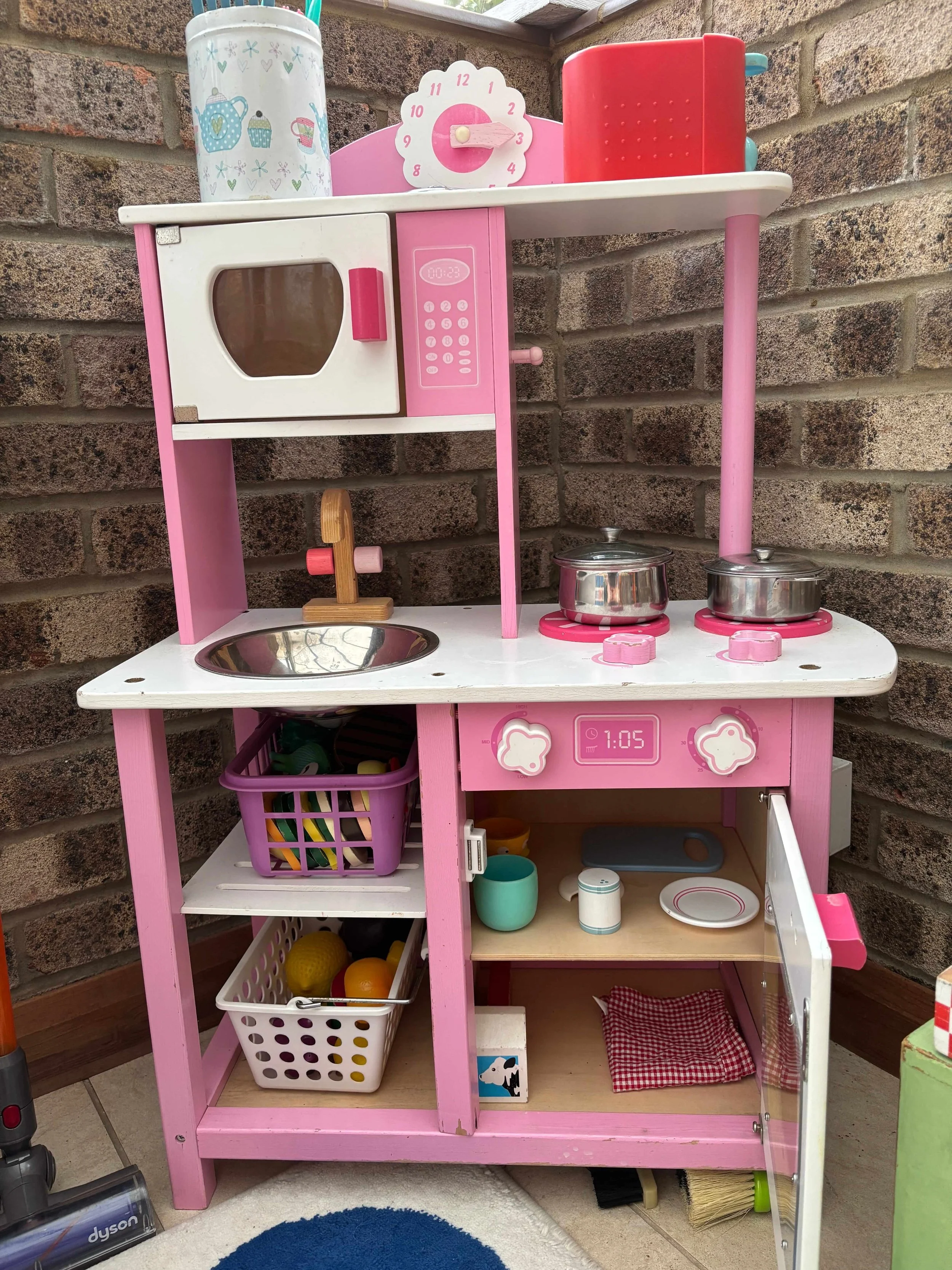 A pink and white toy kitchen set with various toy cooking items and utensils, placed on a tiled floor against a brick wall.