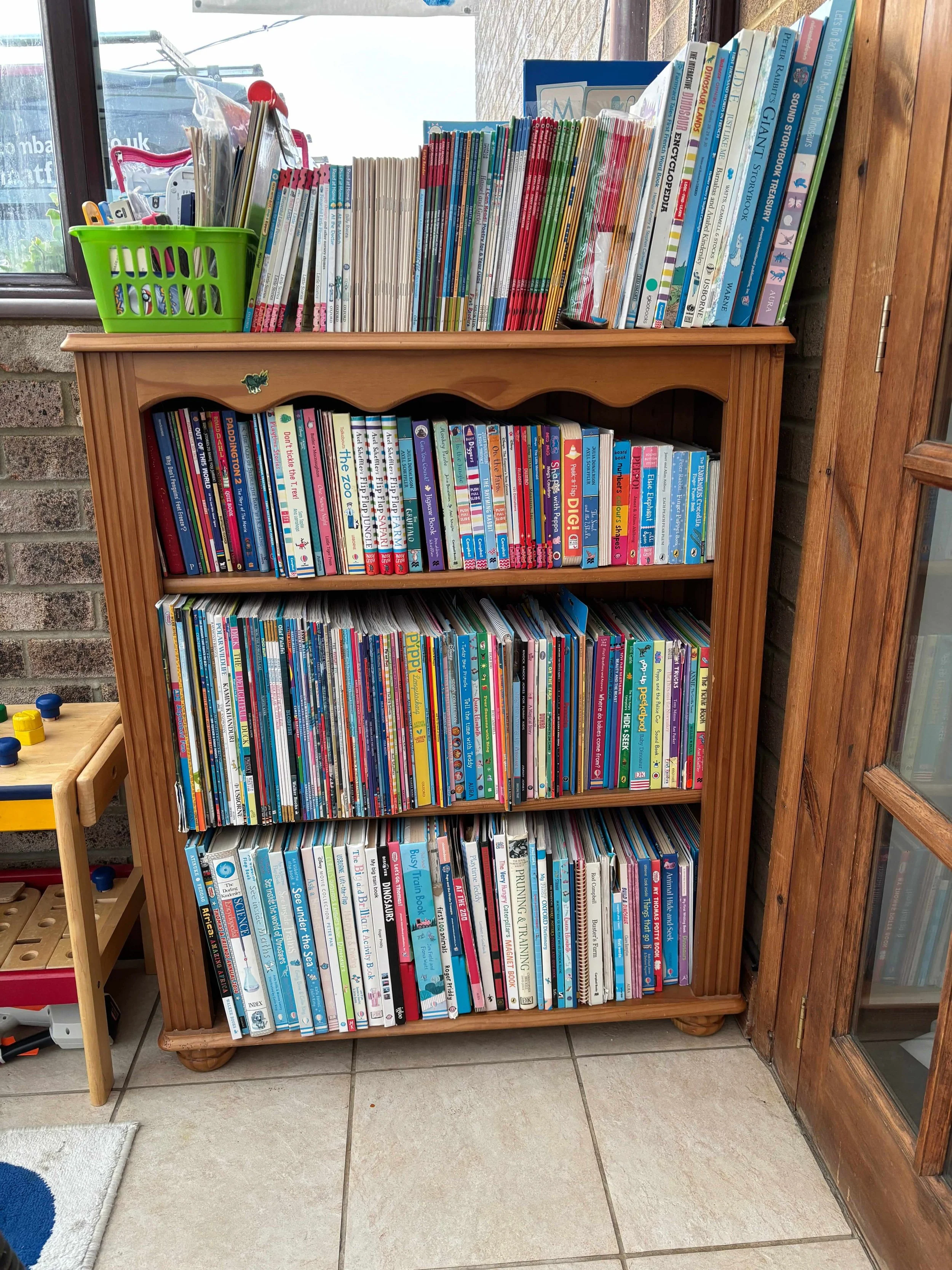 A wooden bookshelf in a children's playroom filled with children's books stored vertically.  There is a green basket on top containing various stationery items, placed next to a window.