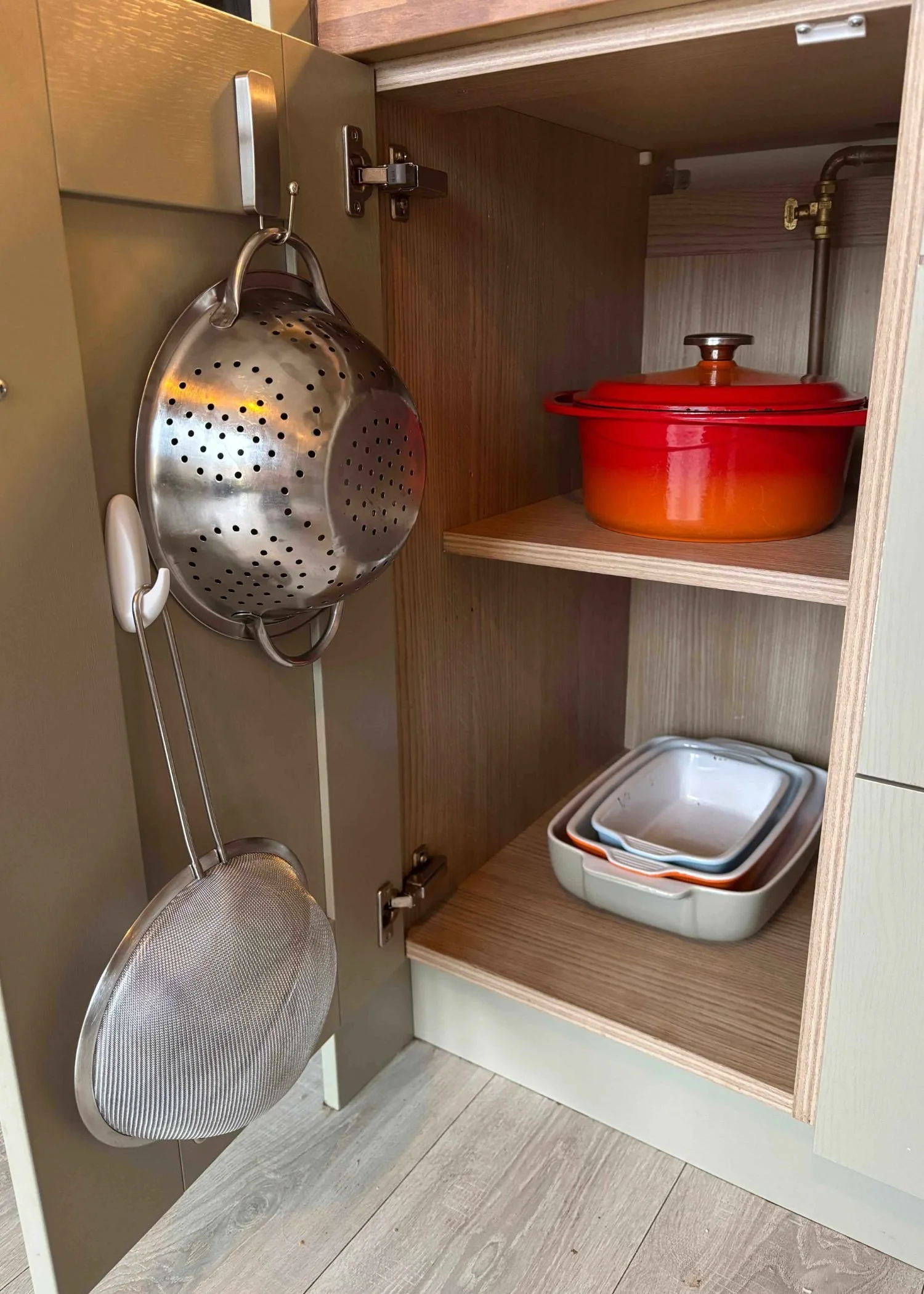An organised kitchen cupboard with an orange le creuset pot, baking dishes and a colander and sieve hanging on the door on hooks.