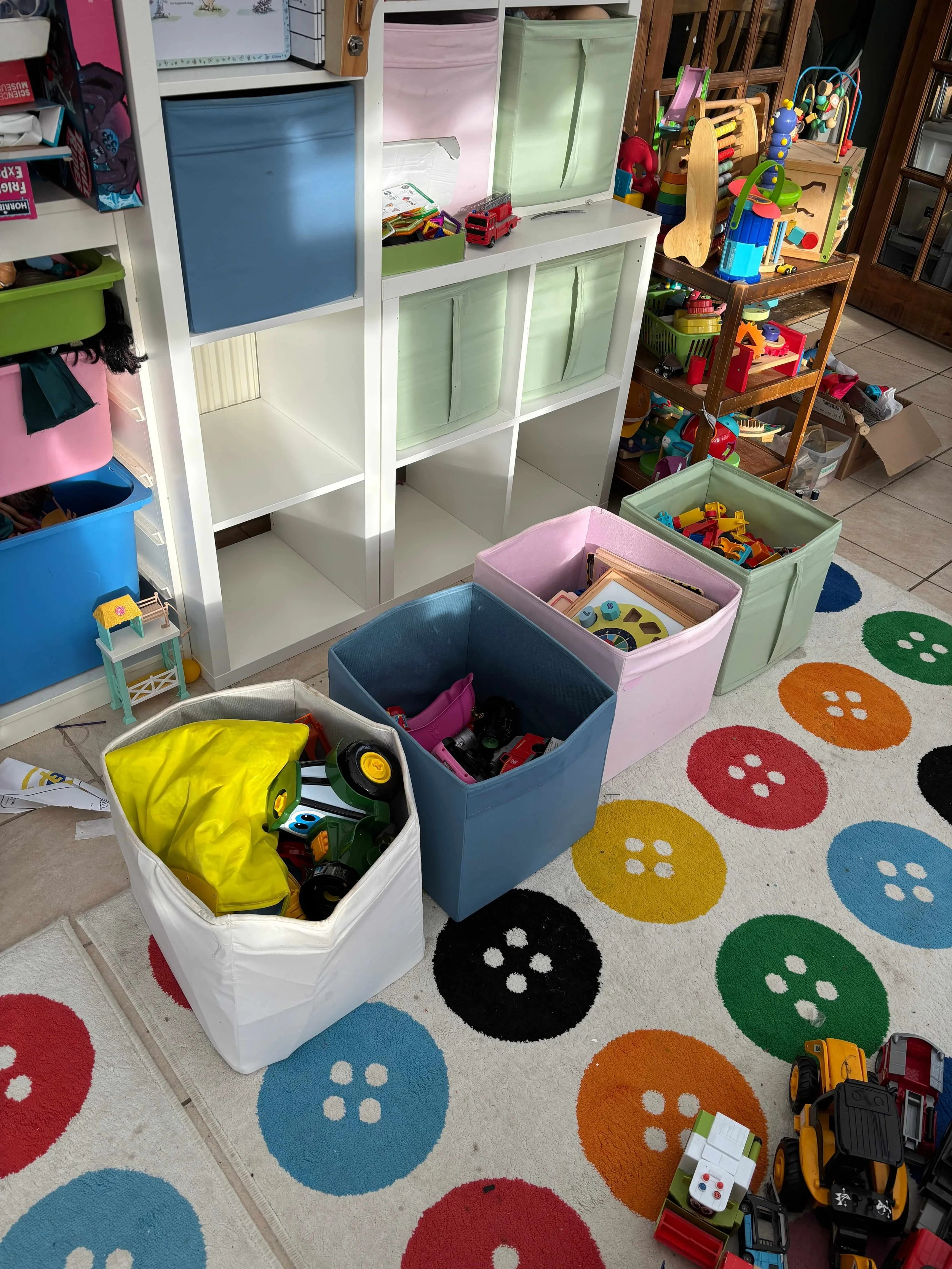 Children's toy storage with bins filled with various toy cars on a colorful, polka-dotted rug.