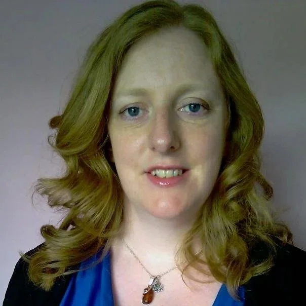A woman with long wavy red hair and blue eyes smiling at the camera, wearing a black top and a blue blouse with a silver necklace featuring a brown stone pendant.