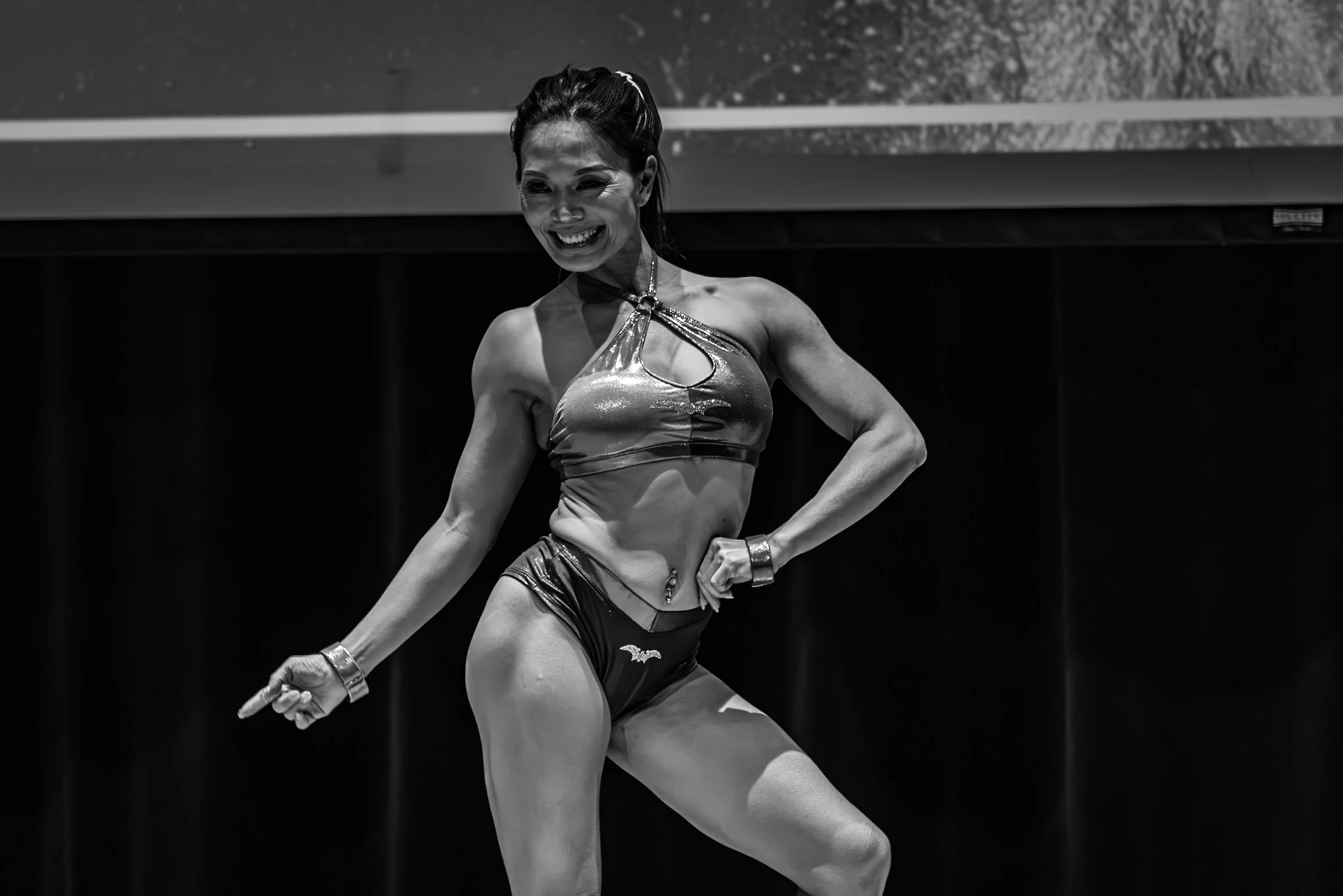 A female bodybuilder posing on stage in a shiny sports bra and shorts, smiling confidently.