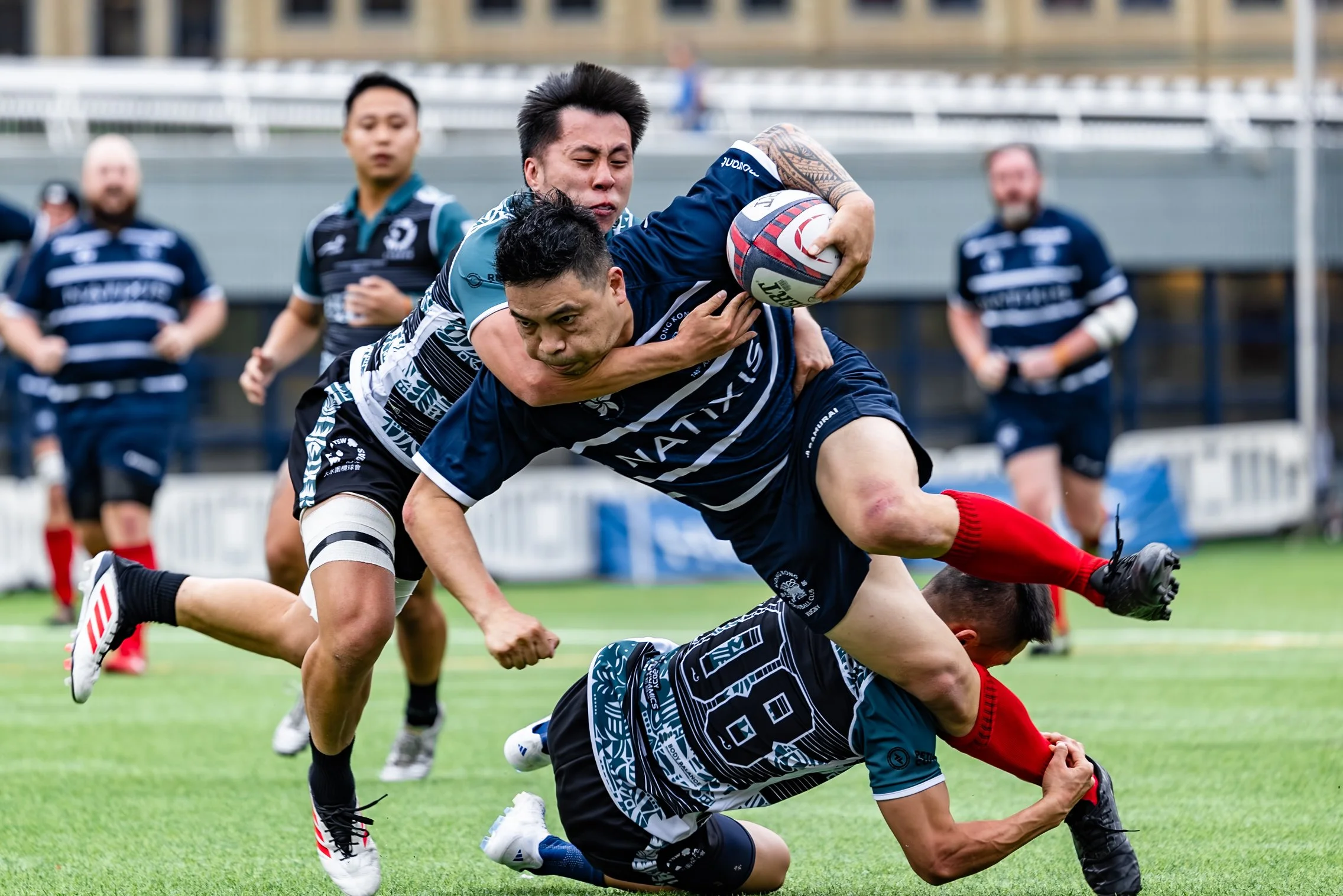 Two rugby players in action during a match, with one player tackling another who is running with the ball. Several other players are visible in the background on the field.