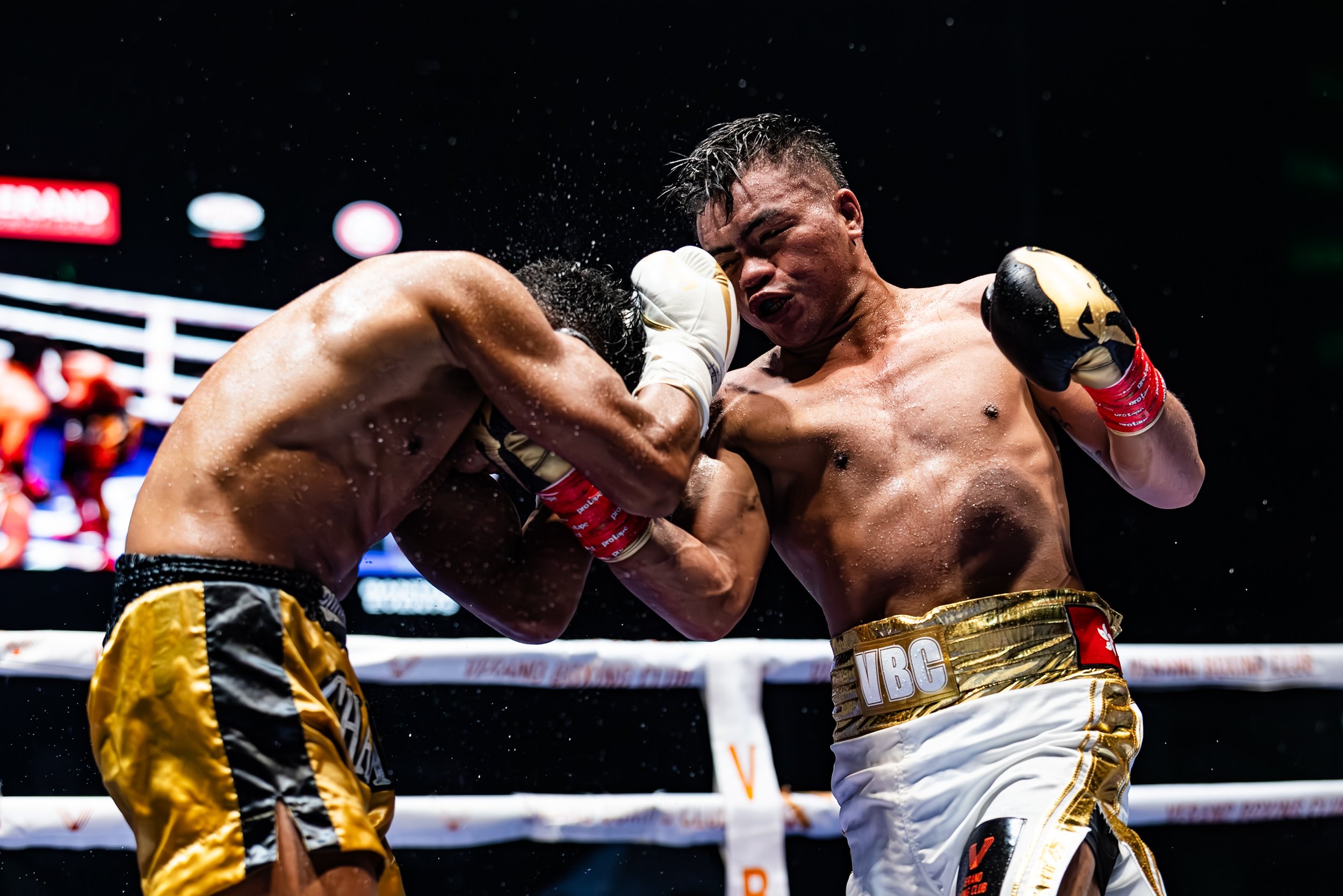 Two shirtless male boxers in a boxing ring engaged in a fight, with one delivering a punch to the other's head. Both are wearing boxing gloves and shorts, and appear sweaty and intense.