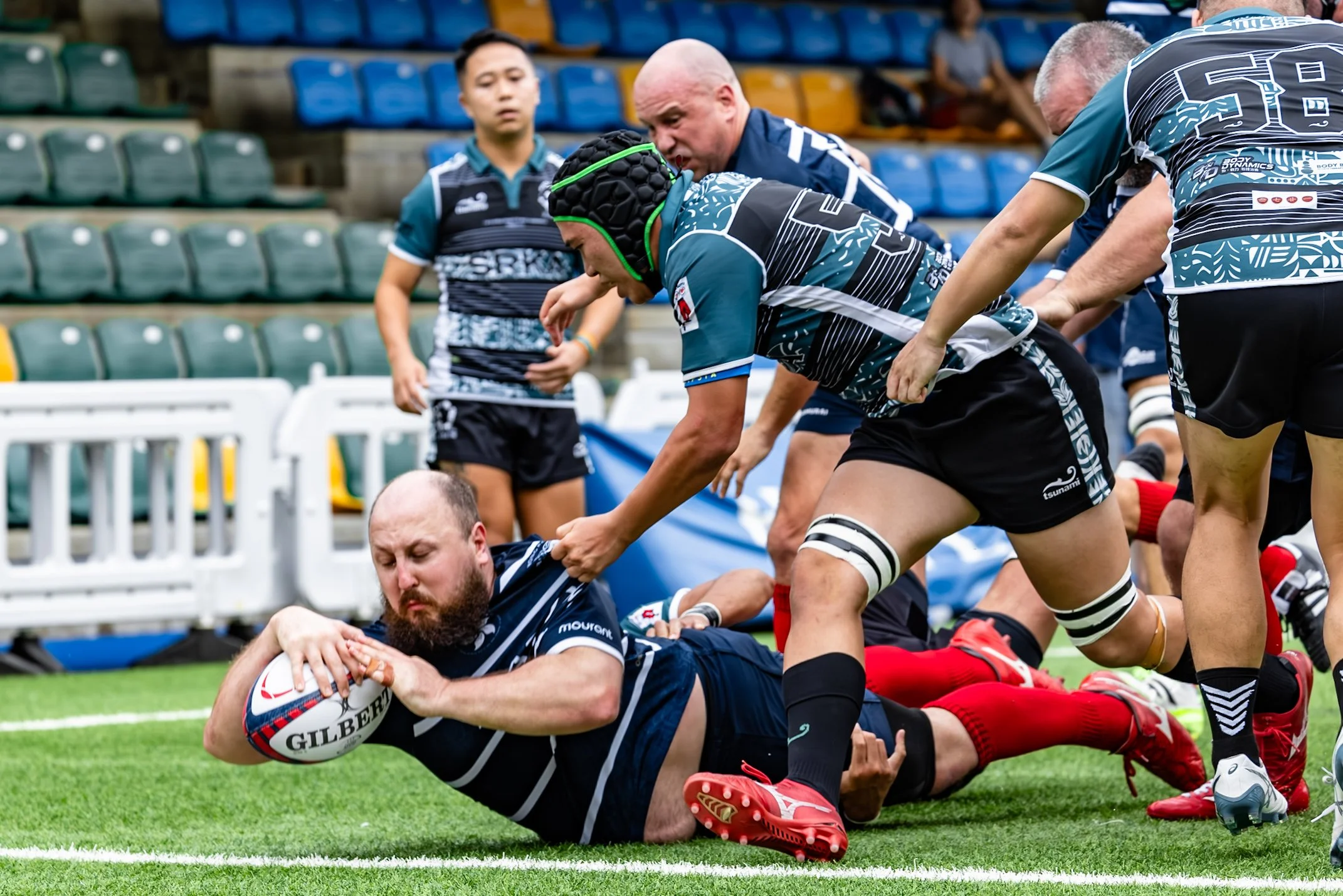 Rugby players competing for control of the ball during a match. Player in navy jersey is on the ground holding the ball, surrounded by teammates and opponents.