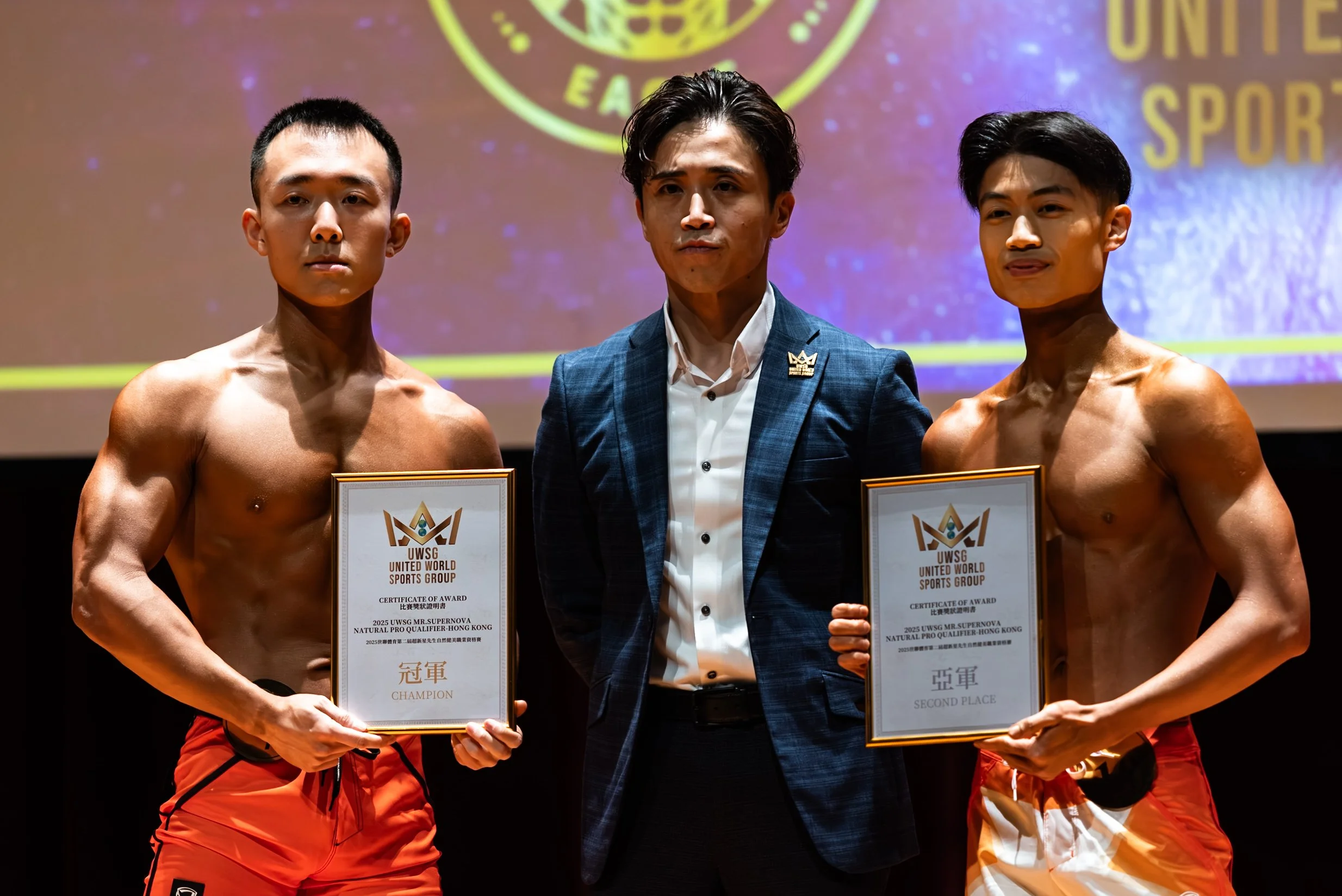 Two shirtless male athletes holding certificates on a stage, standing between a man in a suit, after a sports event. The certificates indicate their achievements in a natural pro qualifier in Hong Kong, with one as champion and the other in second pl