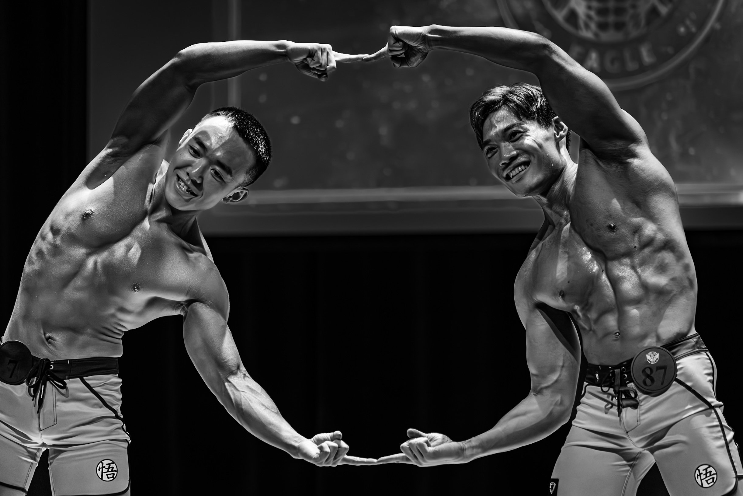 Two shirtless muscular men with short hair and smile, standing on stage, flexing their muscles and touching fists in a celebratory pose, with a dark background and a faint logo.