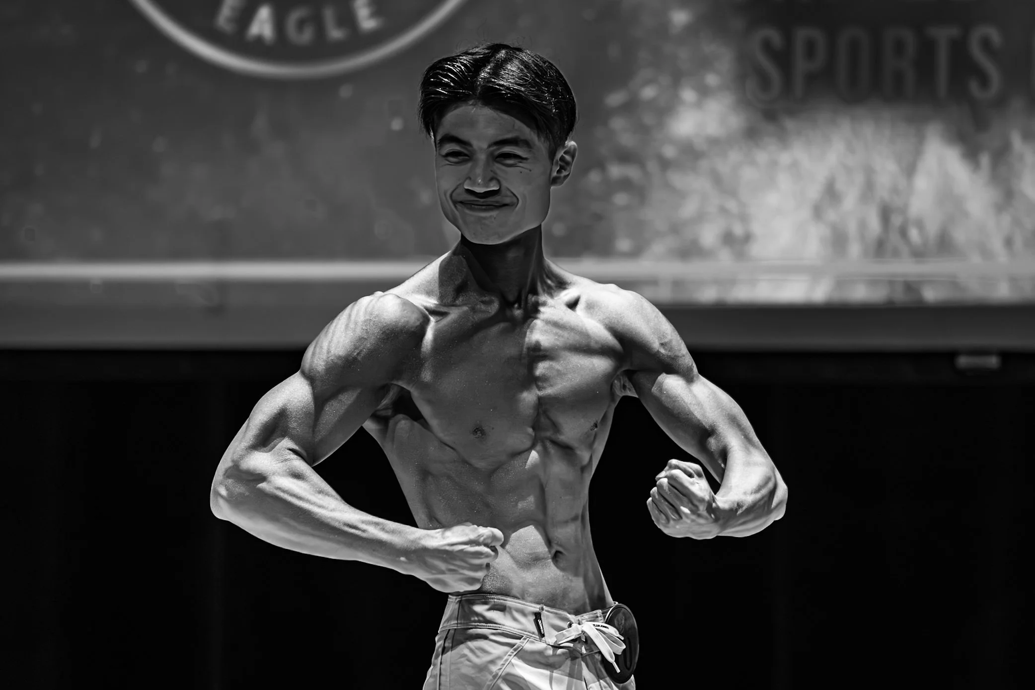 A young man with a muscular physique is posing on stage during a bodybuilding competition, flexing his muscles and smiling. The image is black and white.