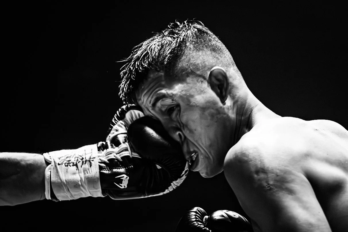 Black and white photo of a boxer with a shaved side hairstyle, grimacing, as he receives a punch to the face.